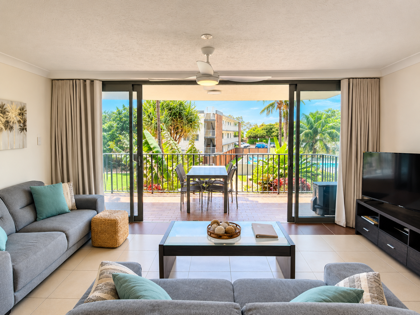 Bright living room with two grey sofas accented by teal and patterned pillows, a glass-topped coffee table with decorative balls and a book, opening through large sliding glass doors to a balcony with an outdoor dining table and four chairs, overlooking lush tropical plants and a neighboring building under a blue sky.