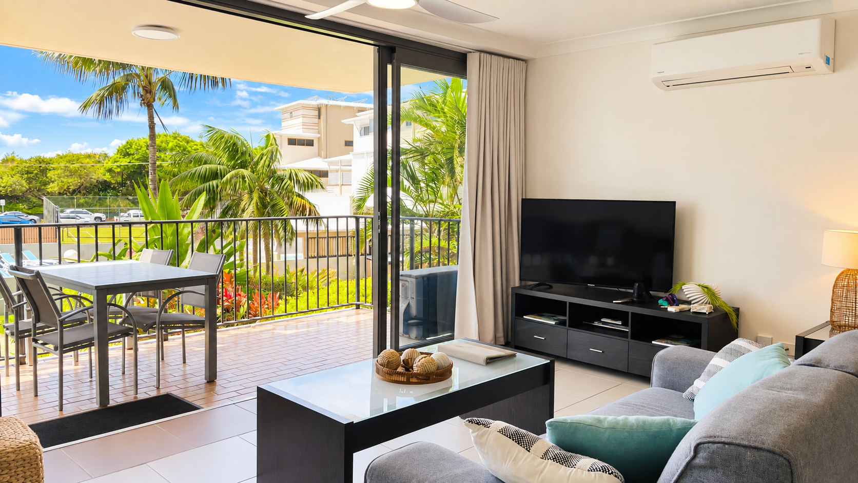 Bright living room with gray sofa adorned with teal and patterned pillows, glass-top coffee table, flat-screen TV on a black stand, sliding glass doors opening to a balcony with outdoor dining table and chairs, overlooking palm trees and a sunny urban view.