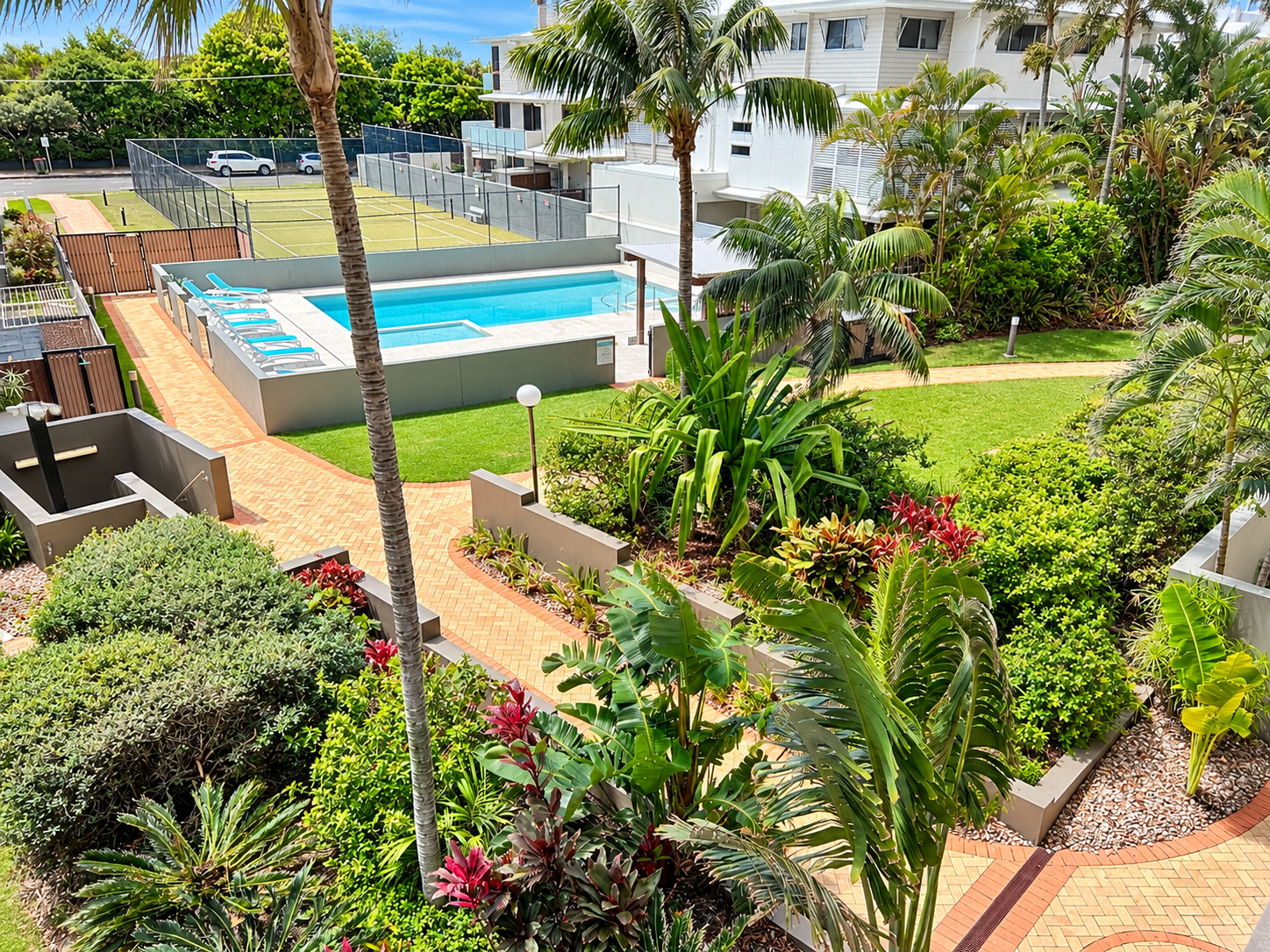 Outdoor view of a residential complex featuring a rectangular swimming pool with lounge chairs, a fenced tennis court, and well-maintained garden paths surrounded by lush green plants and palm trees under a blue sky.
