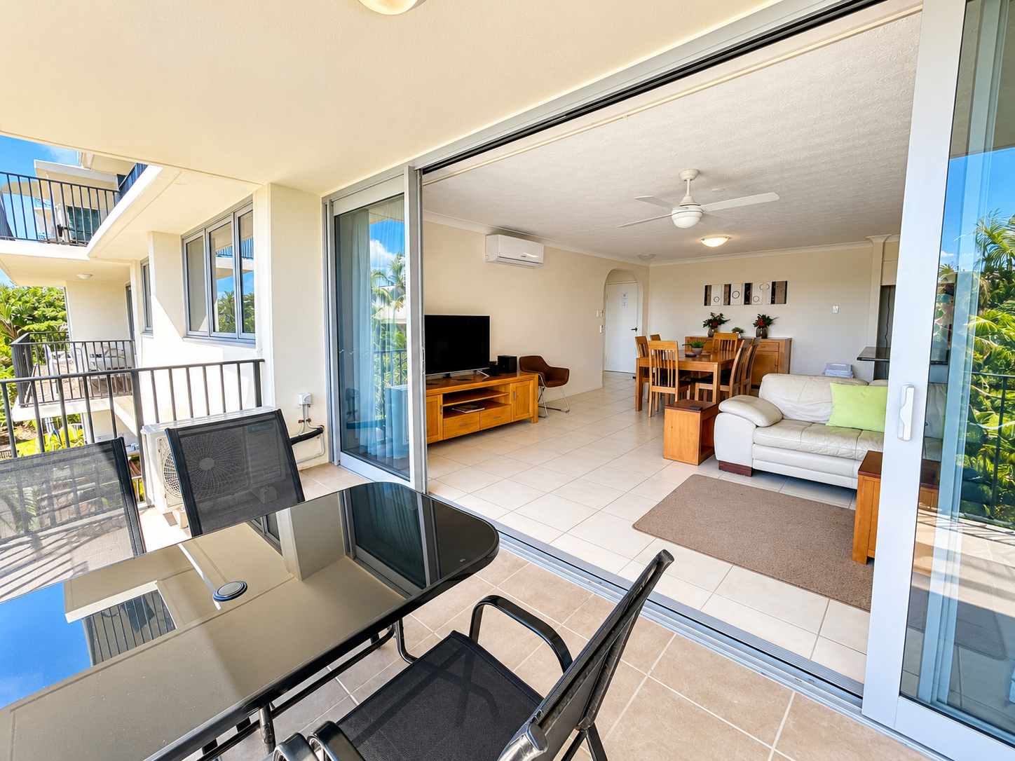 Spacious modern lounge area with a white leather sofa, a wooden dining table with six chairs, a TV on a wooden stand, and a brown chair, opening onto a balcony with a glass-top table and four black chairs overlooking greenery.