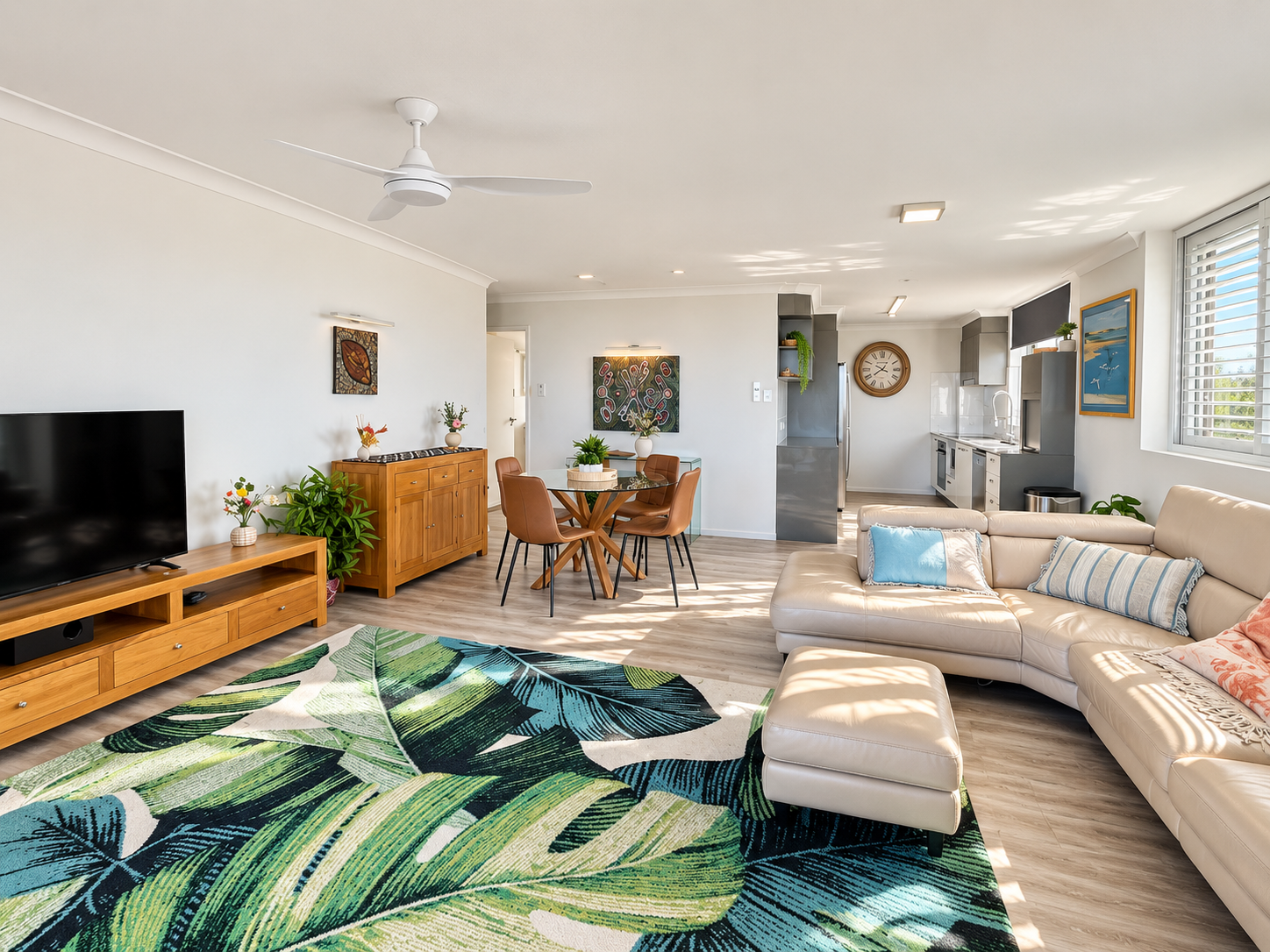 Bright open-plan dining and kitchen area with light wood flooring, a large beige leather sectional sofa with blue and striped cushions, and a matching ottoman. A green and blue leaf-patterned rug covers part of the floor. There is a round glass dining table with four brown chairs, two wooden cabinets with decorative vases and plants, a wall clock, and modern kitchen appliances. Large windows with blinds allow natural light to fill the space.