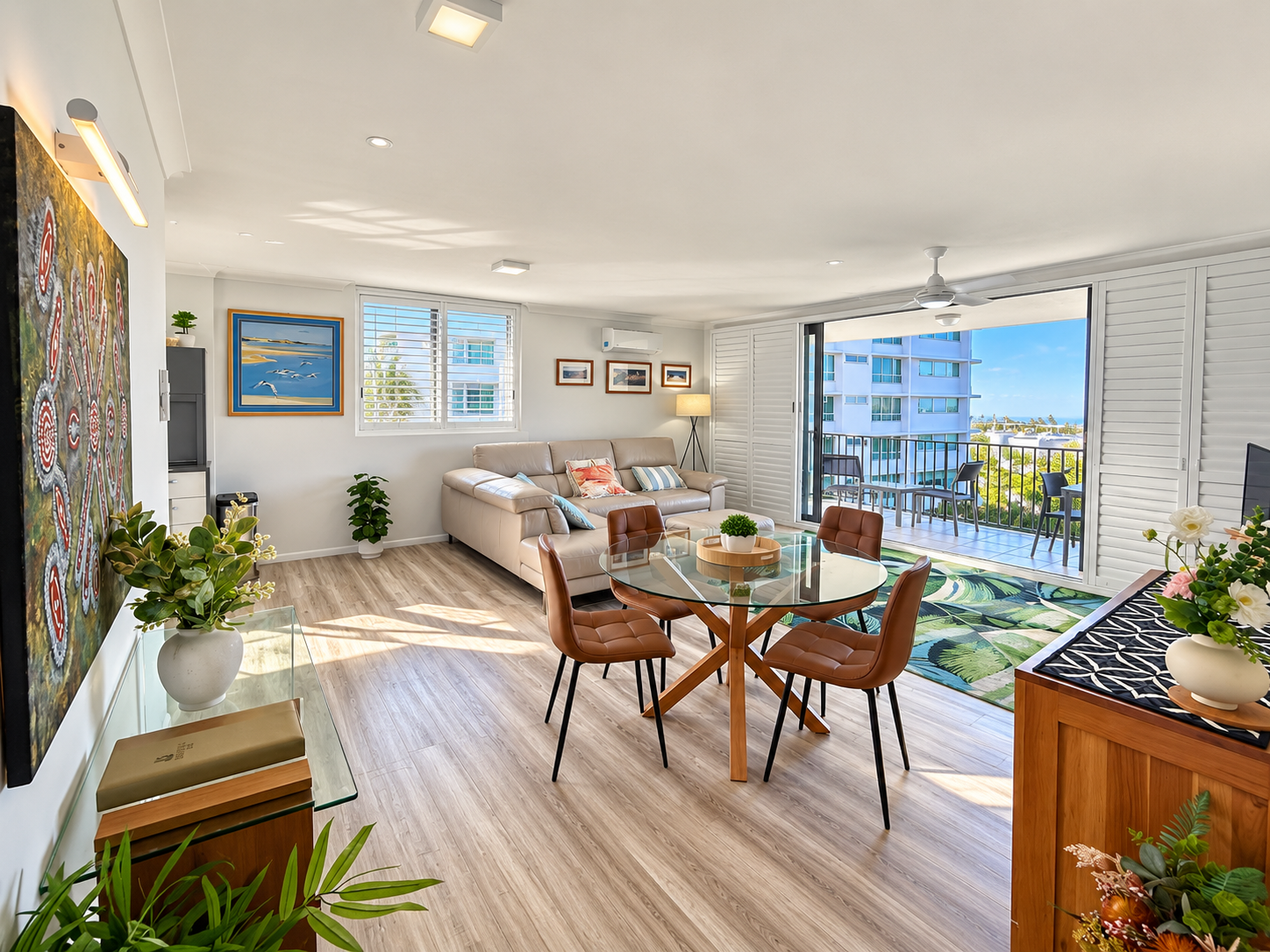 Bright living room with light wood flooring, a beige leather sofa with colorful cushions, a glass-top round dining table with four brown padded chairs, and large open sliding doors leading to a balcony with outdoor chairs and a view of nearby buildings and a blue sky. The room features several framed artworks on the walls, indoor plants, and a ceiling fan.