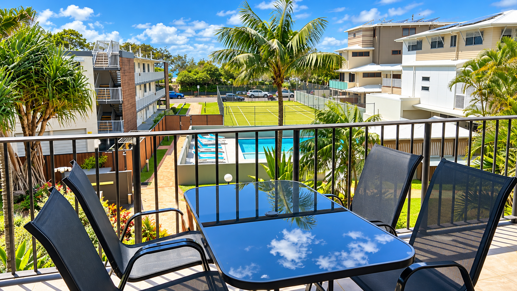 View from a balcony with a black glass-top table and four black mesh chairs overlooking a green tennis court, swimming pool with lounge chairs, palm trees, and modern buildings under a blue sky with scattered clouds.