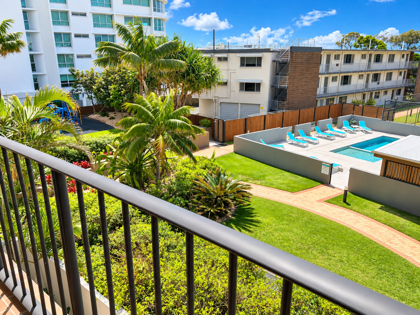 View from a balcony overlooking a swimming pool area with several turquoise lounge chairs arranged along the poolside, surrounded by a gray fence. The pool area is adjacent to a grassy lawn with a brick walkway cutting through it. Tall palm trees and lush green bushes are visible near the balcony and throughout the courtyard. In the background, there are multiple apartment buildings under a bright blue sky with scattered white clouds.