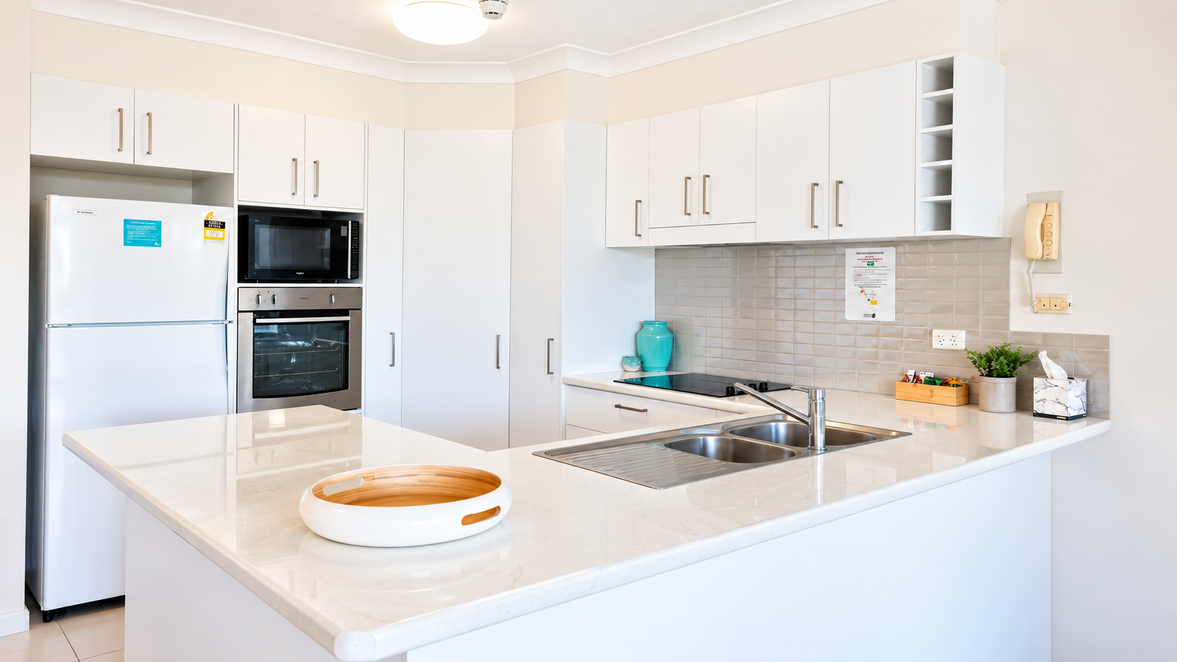 Modern kitchen with white cabinetry and beige tile backsplash, featuring a stainless steel microwave, oven, and refrigerator; a white countertop island with a round wooden tray and a double sink with a chrome faucet; small decorations on the counter include a green plant, tissue box, turquoise vase, and condiment box.