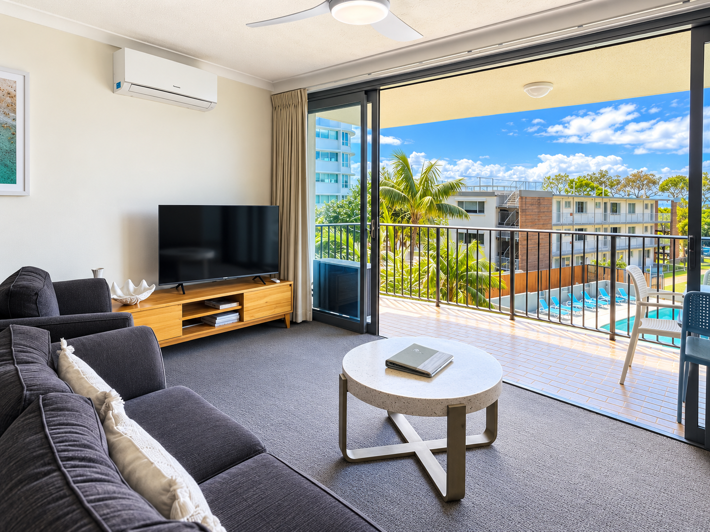 Modern living room with dark gray sofa and armchair, white round coffee table, flat-screen TV on wooden stand, and sliding glass doors opening to a balcony overlooking palm trees, a pool with blue lounge chairs, and nearby buildings under a blue sky with scattered clouds.