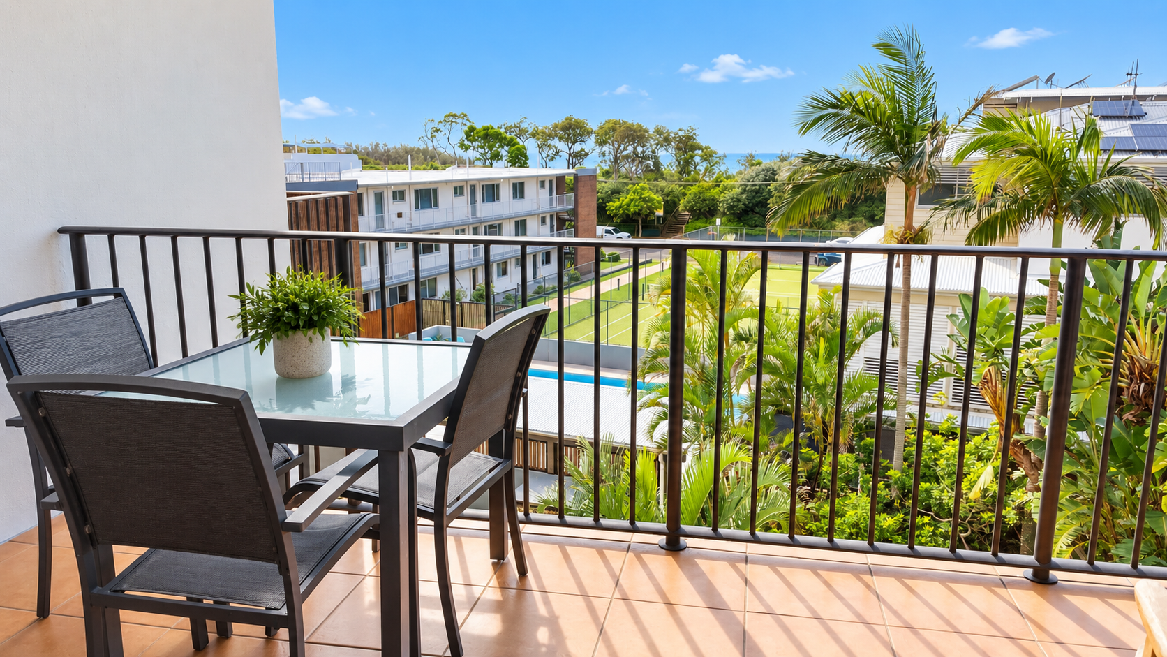 Balcony with a square glass-top table and three black metal chairs, a small potted plant on the table, overlooking a lush green garden, palm trees, a tennis court, and nearby buildings under a bright blue sky.
