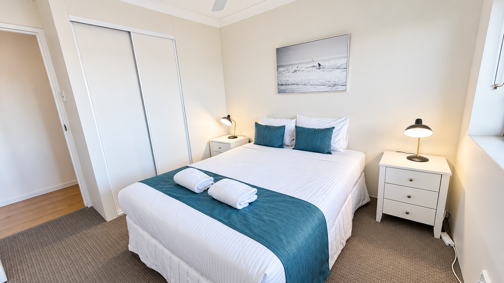 Bright, tidy bedroom with a double bed featuring white linens, teal bed runner and matching pillows, two rolled white towels on the bed, bedside tables with lamps, a ceiling fan, a closet with sliding doors, beige walls, carpeted floor, and a framed black and white photograph above the bed.