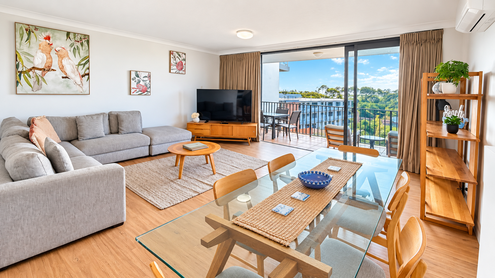 Bright living room with a light gray L-shaped sofa, round wooden coffee table, large flat-screen TV on wooden stand, glass dining table with wooden chairs, open shelves with plants and decor, and sliding glass doors opening to a balcony with outdoor seating and a view of trees and buildings under a blue sky.