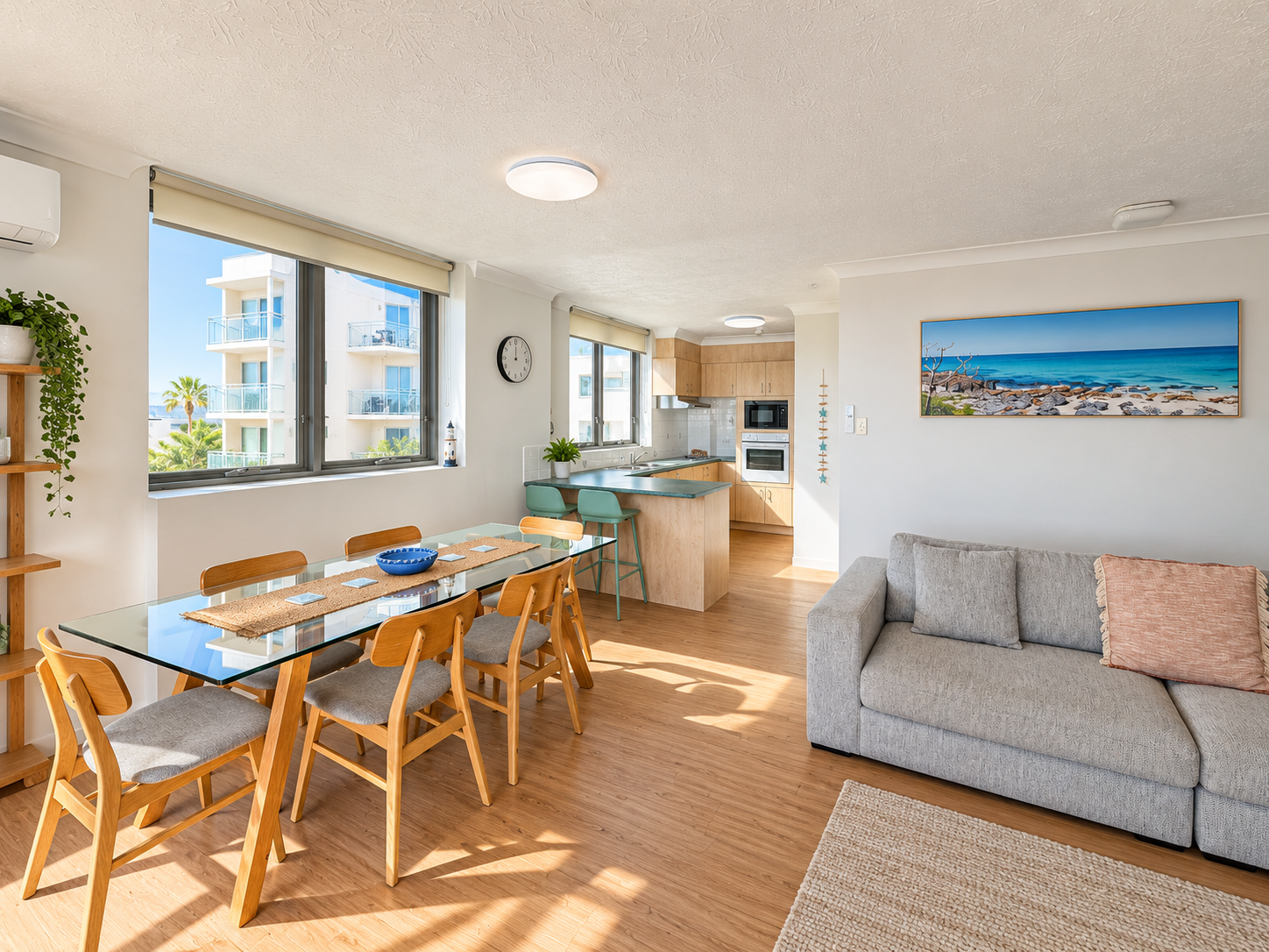 Bright open-plan living space with a glass dining table with six wooden chairs on the left, a kitchen with wood cabinets and green counter stools in the background, and a gray sofa with cushions on the right under a framed ocean landscape painting.