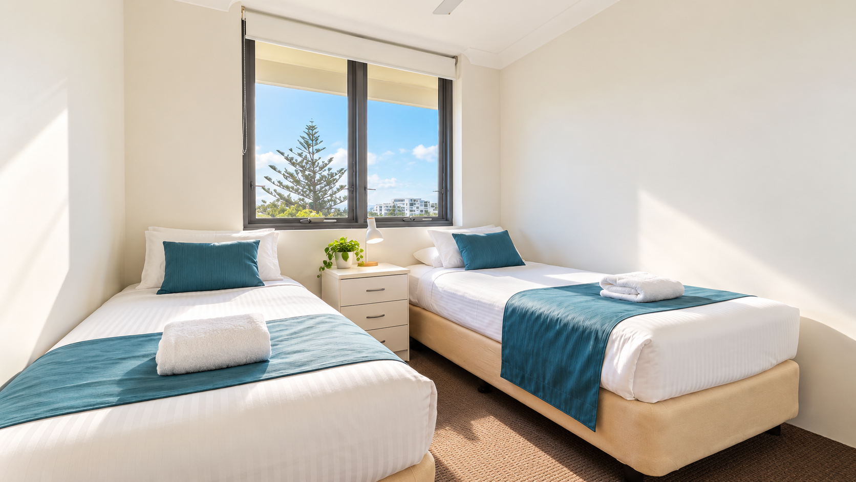 Bright twin bedroom with two single beds featuring white linens, teal runners and pillows, a folded white towel on each bed, a small bedside table with a lamp and a green plant, and a large window showing blue sky and trees outside.
