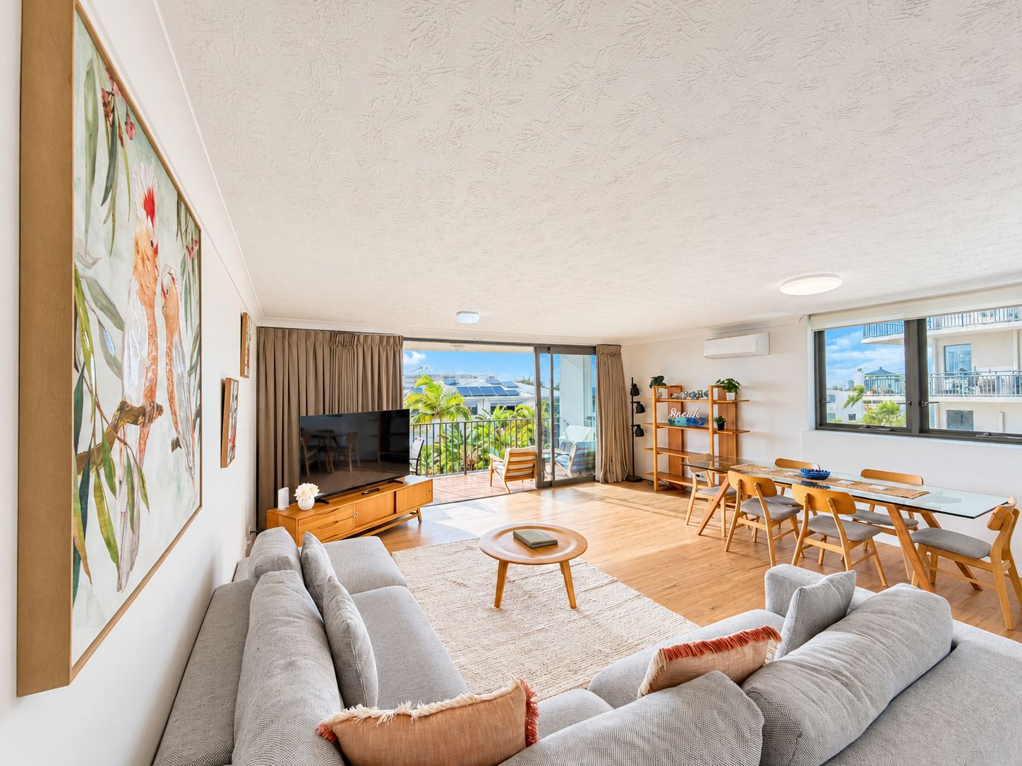Spacious, bright living and dining area with light wood flooring, a large gray sectional sofa with orange pillows, a round wooden coffee table, and a TV on a wooden stand. The room opens to a balcony with outdoor chairs and tropical plants visible outside. There is a wooden dining table with six chairs next to a large window, and a wooden shelving unit with decor and plants under an air conditioner.