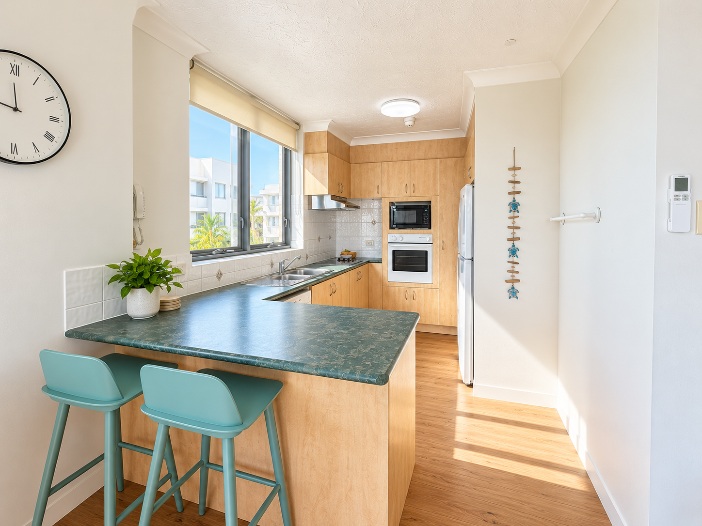 Bright kitchen with light wood cabinets, green marble-like countertop, two teal bar stools, double sink under large windows, built-in oven and microwave, white refrigerator, wall clock with Roman numerals, and sunlight casting shadows on the wooden floor.