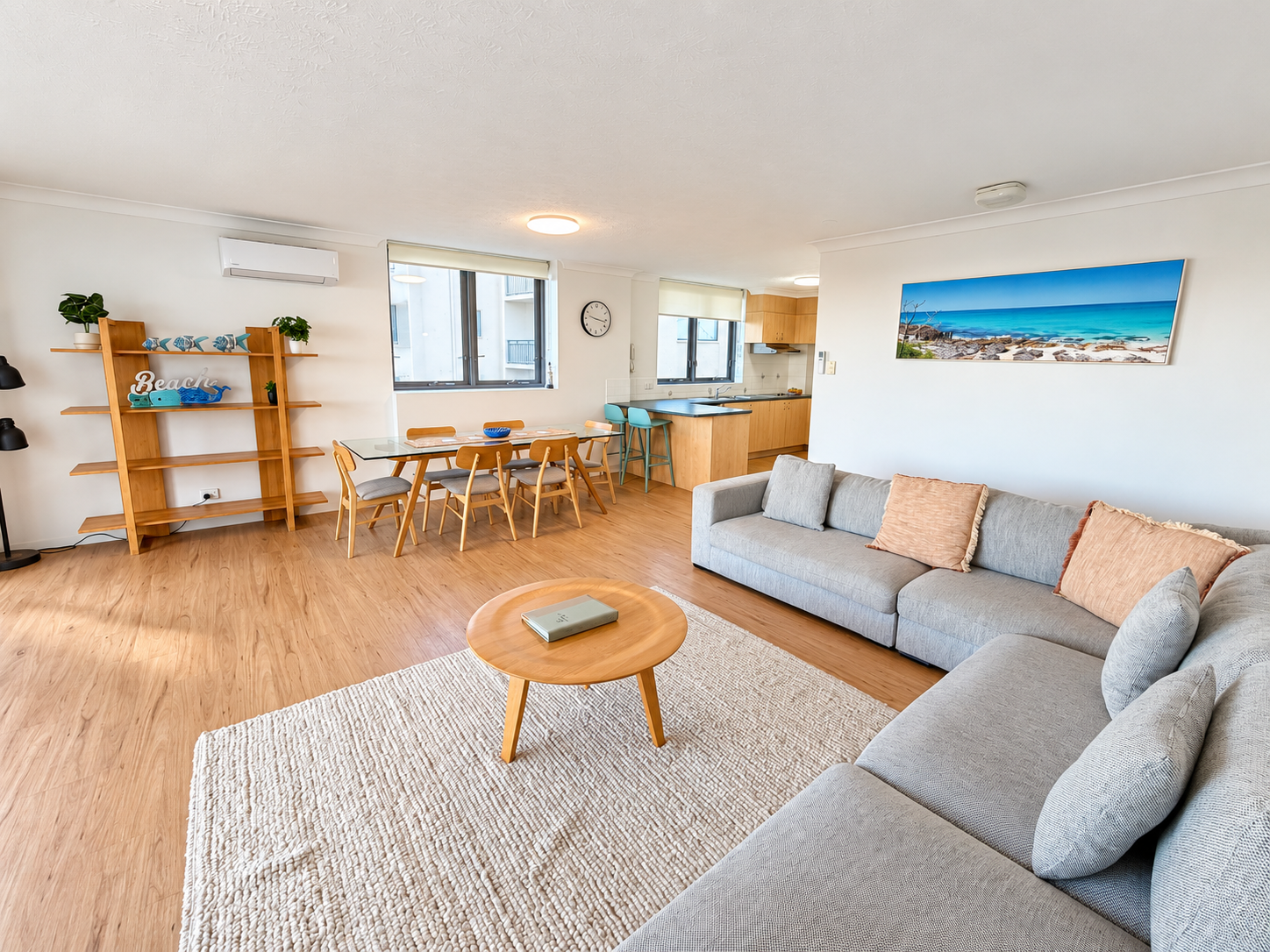 Bright living area with a large gray sectional sofa with peach and gray cushions, a round light wood coffee table with a book, a textured white rug, a glass dining table with six wooden chairs, and an open kitchen with wood cabinetry and two turquoise bar stools. A wooden shelf on the left holds plants and decorative items including a 'Beach' sign. A panoramic beach photo hangs on the wall above the sofa. Two windows provide natural light.