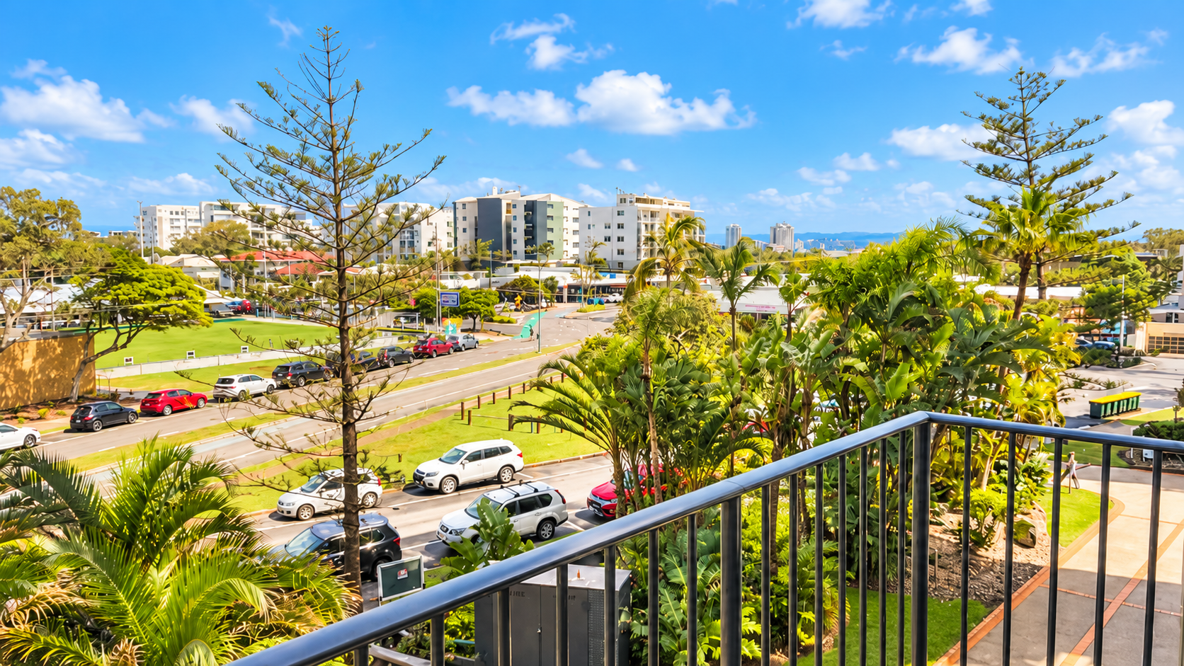 View from a balcony with metal railing overlooking a street lined with parked cars, lush green tropical plants, trees, and residential buildings under a bright blue sky with scattered clouds.