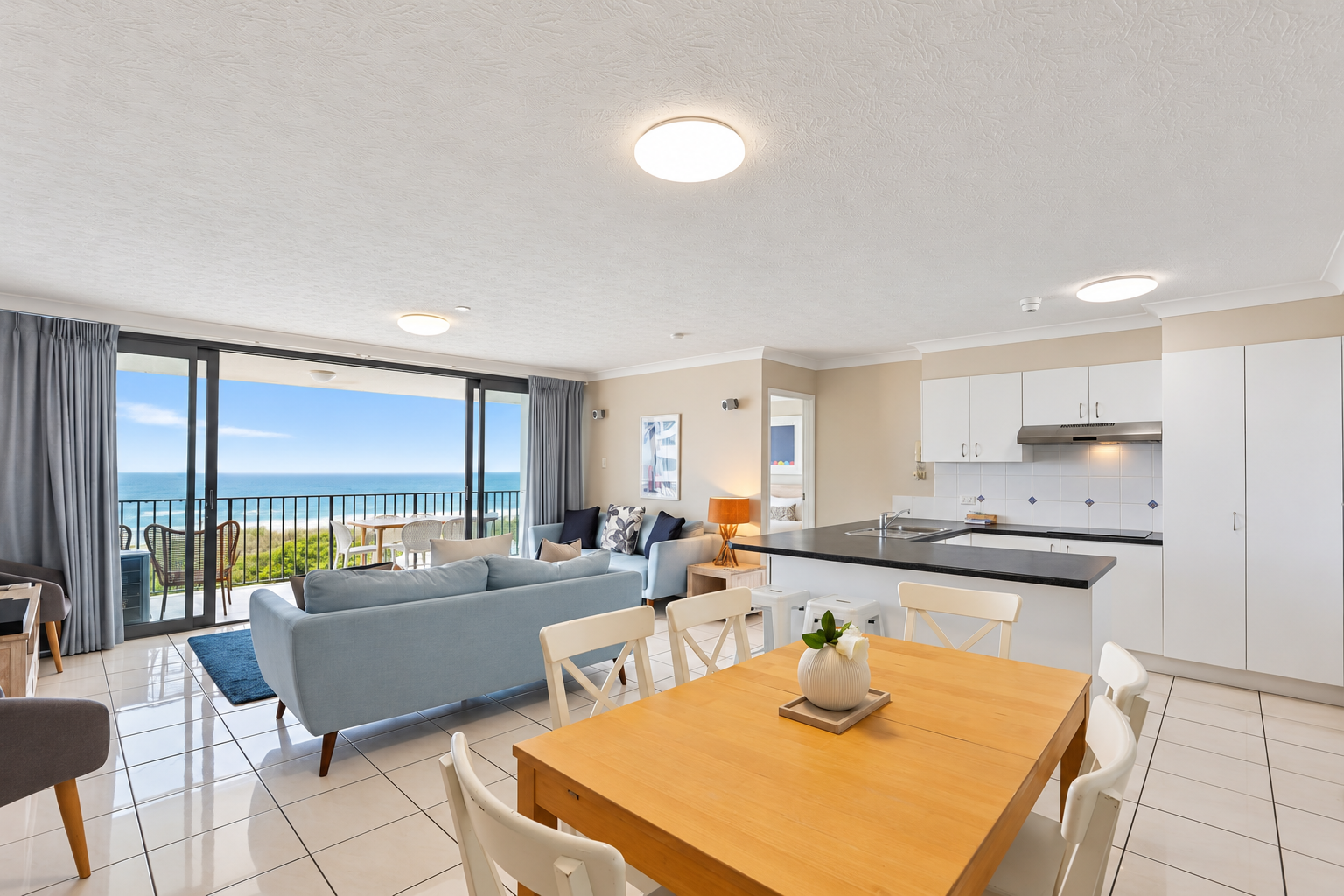 Bright, modern living area with light blue sofas facing a sliding glass door that opens to a balcony with outdoor seating and a view of the ocean and beach, adjacent to an open kitchen with white cabinets and a black countertop, and a wooden dining table with white chairs and a small white vase with a flower.