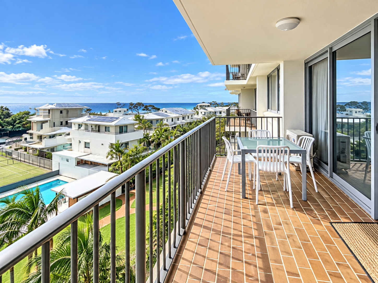 Balcony with brown tiled floor, glass table, and white chairs overlooking landscaped gardens, swimming pool, tennis court, white residential buildings, and ocean under a partly cloudy blue sky.
