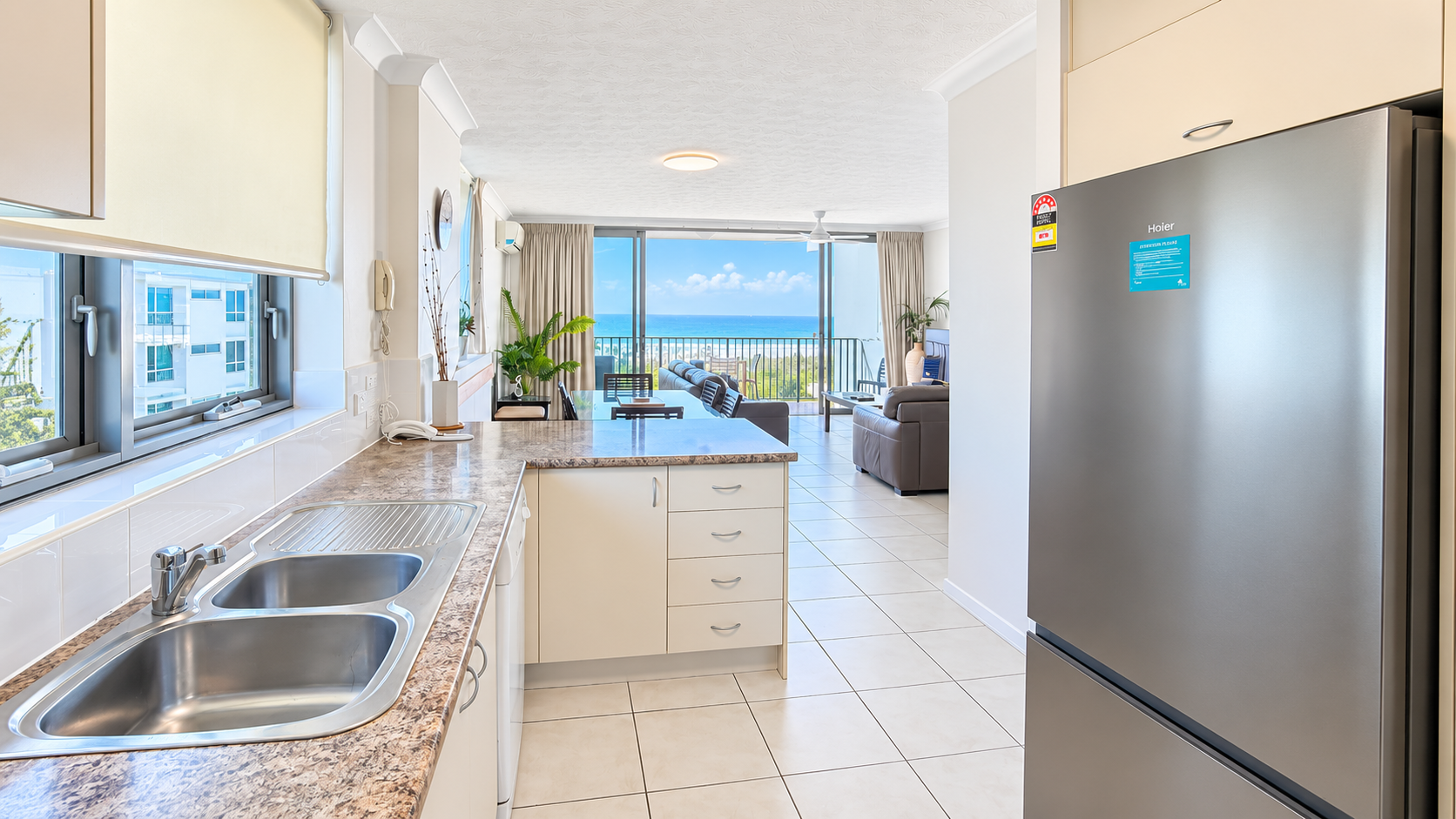 Modern kitchen with beige countertops and a double stainless steel sink on the left, a large stainless steel refrigerator on the right, and an open view into a living area with a sofa and large windows overlooking a bright blue ocean under a partly cloudy sky.