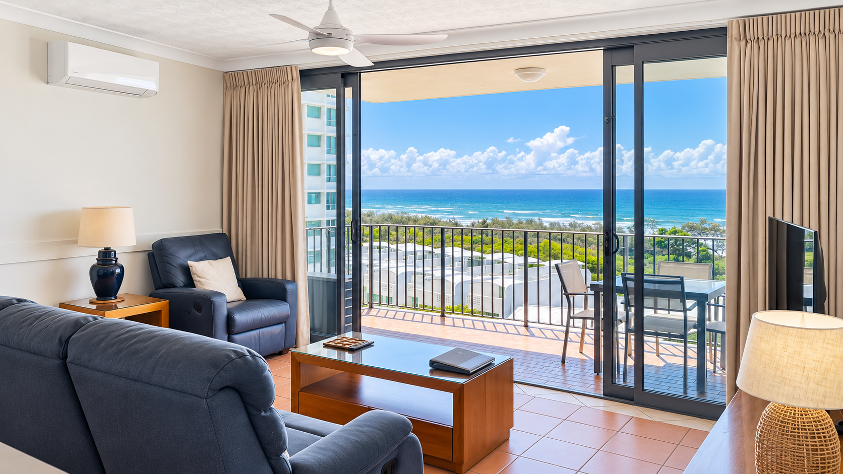 Modern living room with blue leather sofa and armchair, wooden coffee table, two lamps, and large sliding glass doors opening to a balcony overlooking the ocean with blue sky and scattered clouds.