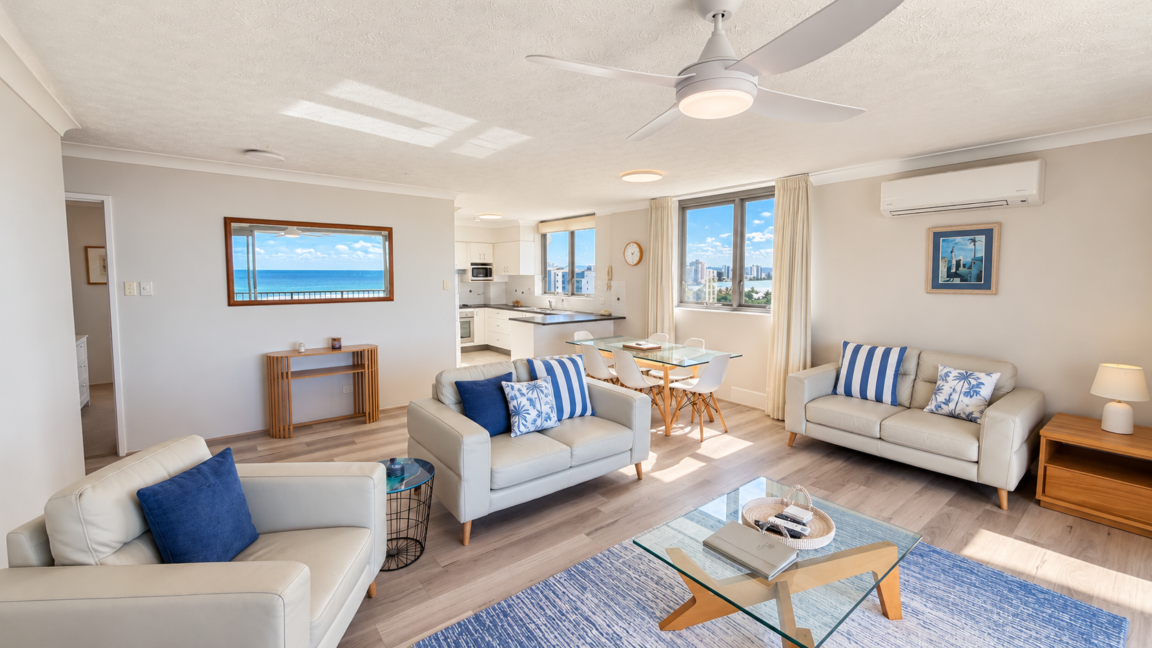 Bright living room with beige leather sofas adorned with blue and white pillows, a glass coffee table on a blue rug, dining area with glass table and white chairs, modern kitchen in the background, large windows with views of the ocean and city skyline, wooden side tables, ceiling fan, and a framed painting on the wall.