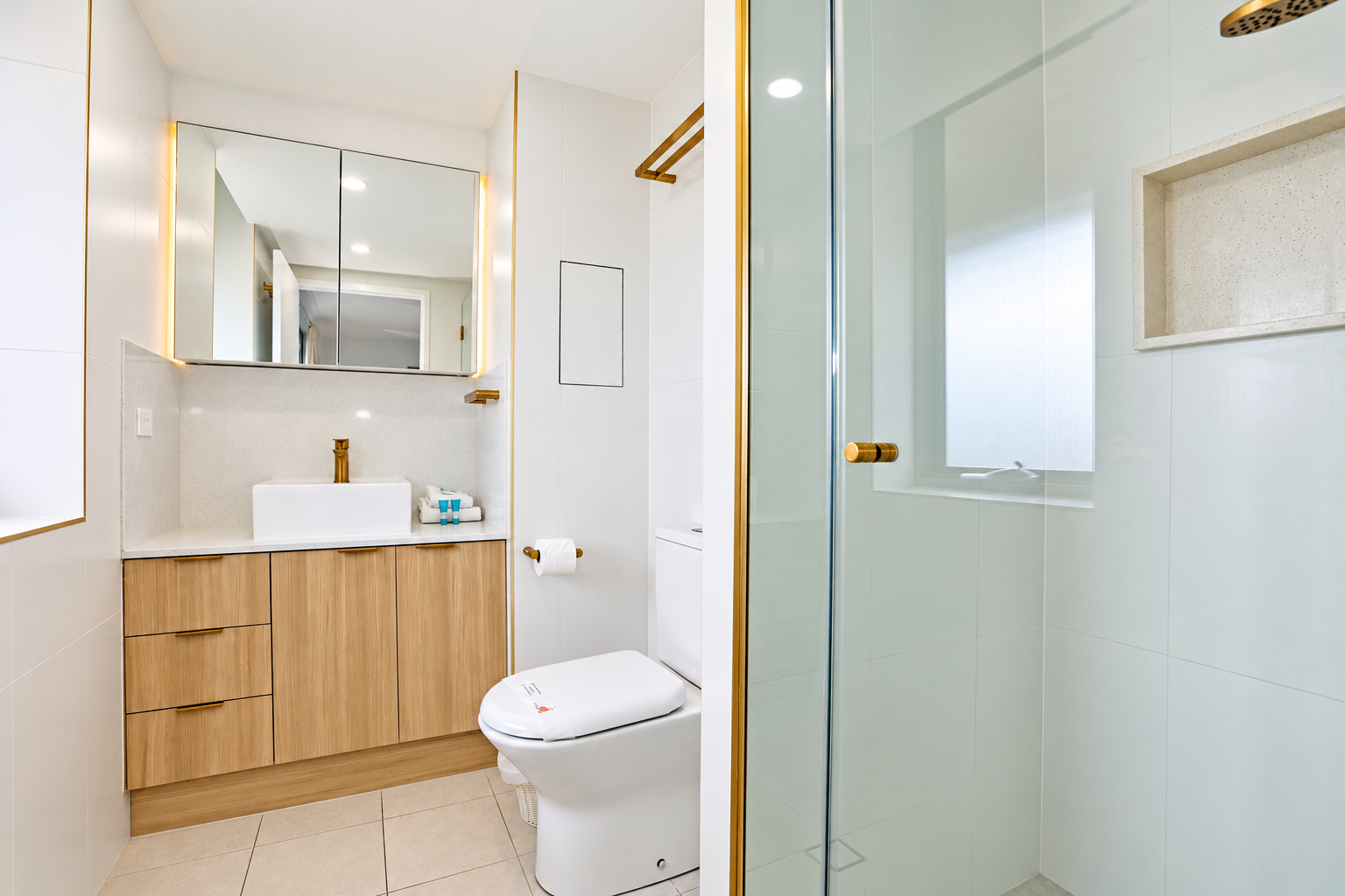 Modern bathroom featuring a white toilet with a seat cover, wooden vanity with a white rectangular sink and gold faucet, illuminated mirrored cabinet above, and a glass shower enclosure with gold hardware and a built-in shelf near a frosted window.