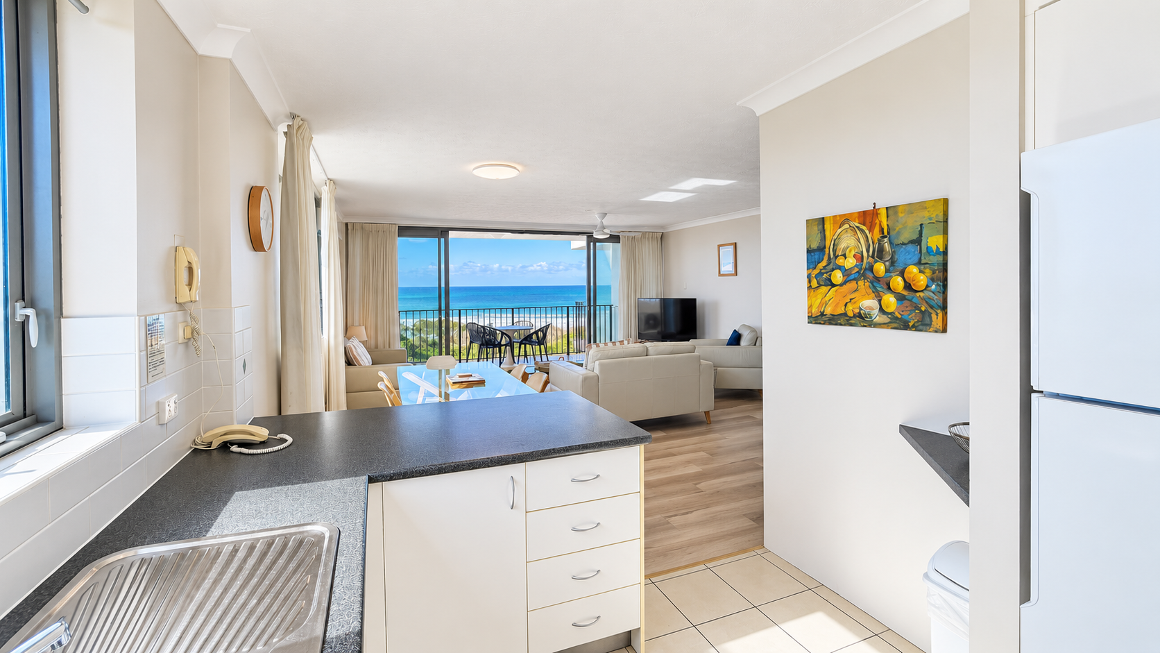 Bright kitchen with black countertop, white cabinets, and a stainless steel sink leading to a living and dining area with beige sofas, a glass table with chairs, a TV, and floor-to-ceiling windows showing a balcony overlooking a blue ocean and sky.
