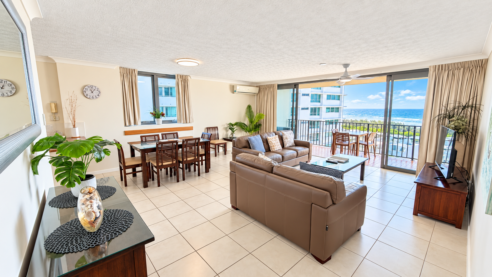 Bright living and dining area featuring beige tiled floors, two tan leather couches with patterned cushions around a glass coffee table, a wooden dining table with six chairs, indoor plants, and large glass sliding doors opening to a balcony with ocean views.