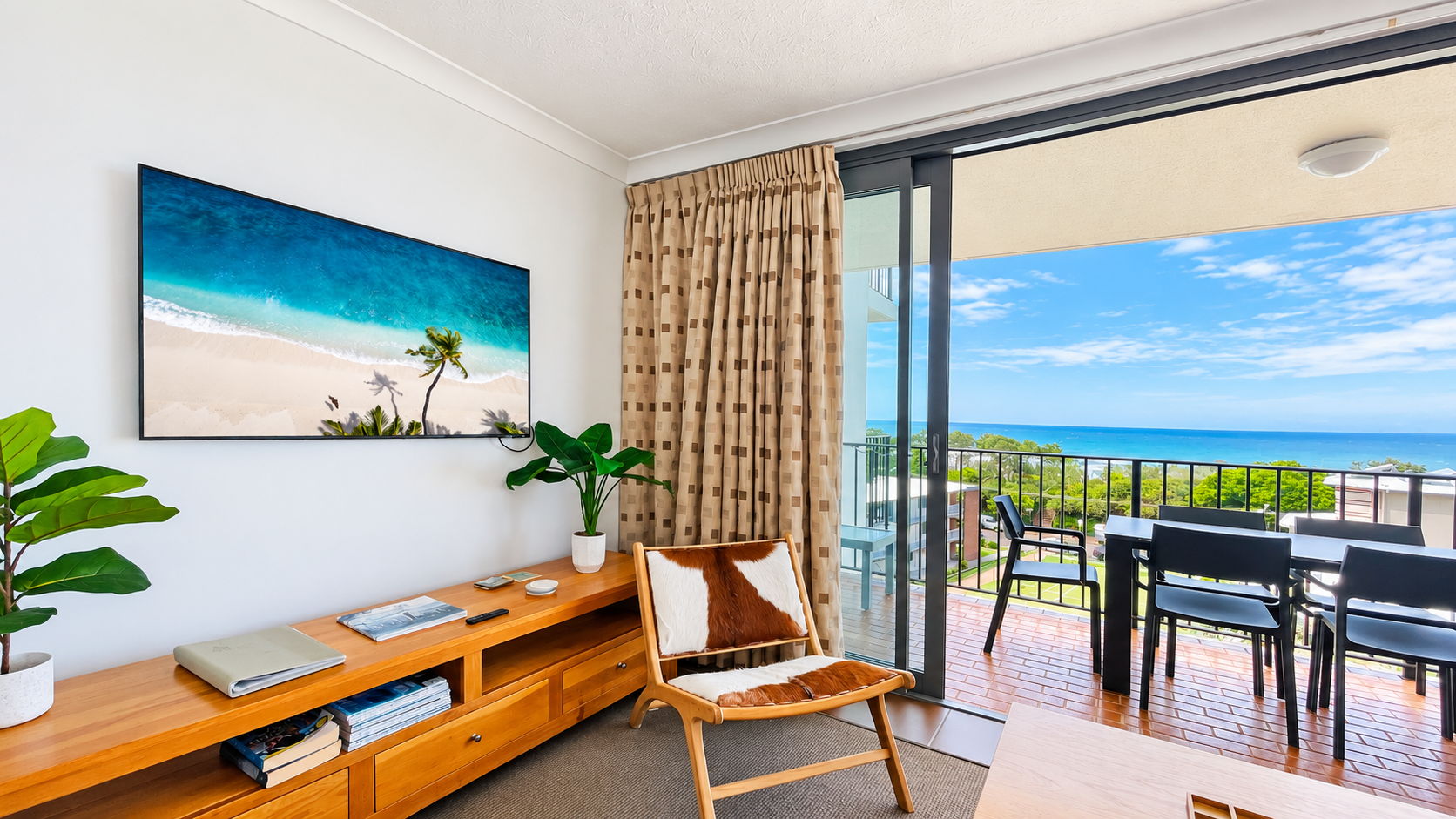 Modern lounge area with a wooden chair featuring brown and white cowhide upholstery, a wooden TV stand with books and plants, a wall-mounted flat-screen TV showing a beach scene, and sliding glass doors opening to a balcony with black chairs and table overlooking a blue ocean and green trees under a partly cloudy sky.