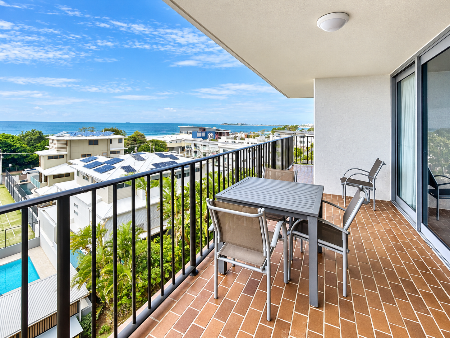 Spacious balcony with terracotta tile flooring, a table and four chairs, overlooking a coastal neighborhood with houses, palm trees, a swimming pool, and the ocean under a partly cloudy blue sky.