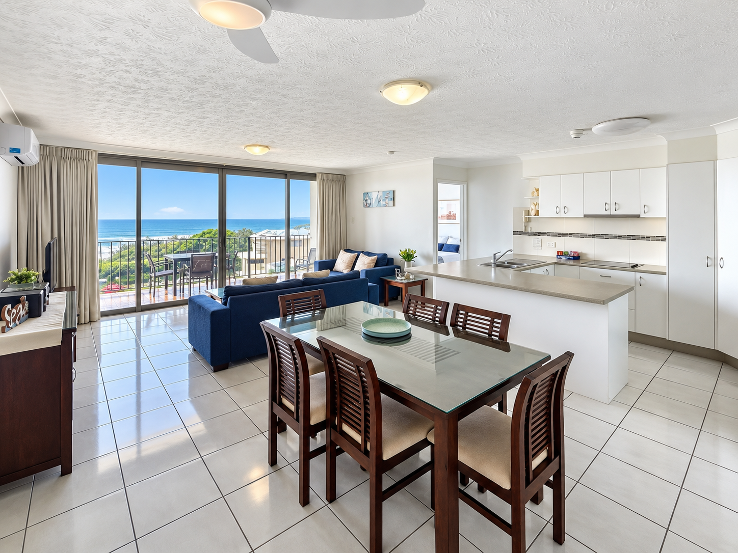 Open plan dining and kitchen area with a glass-top dining table surrounded by six wooden chairs, a kitchen island with a sink, white cabinetry, and a living space with blue sofas facing a large sliding glass door that opens to a balcony with ocean views.