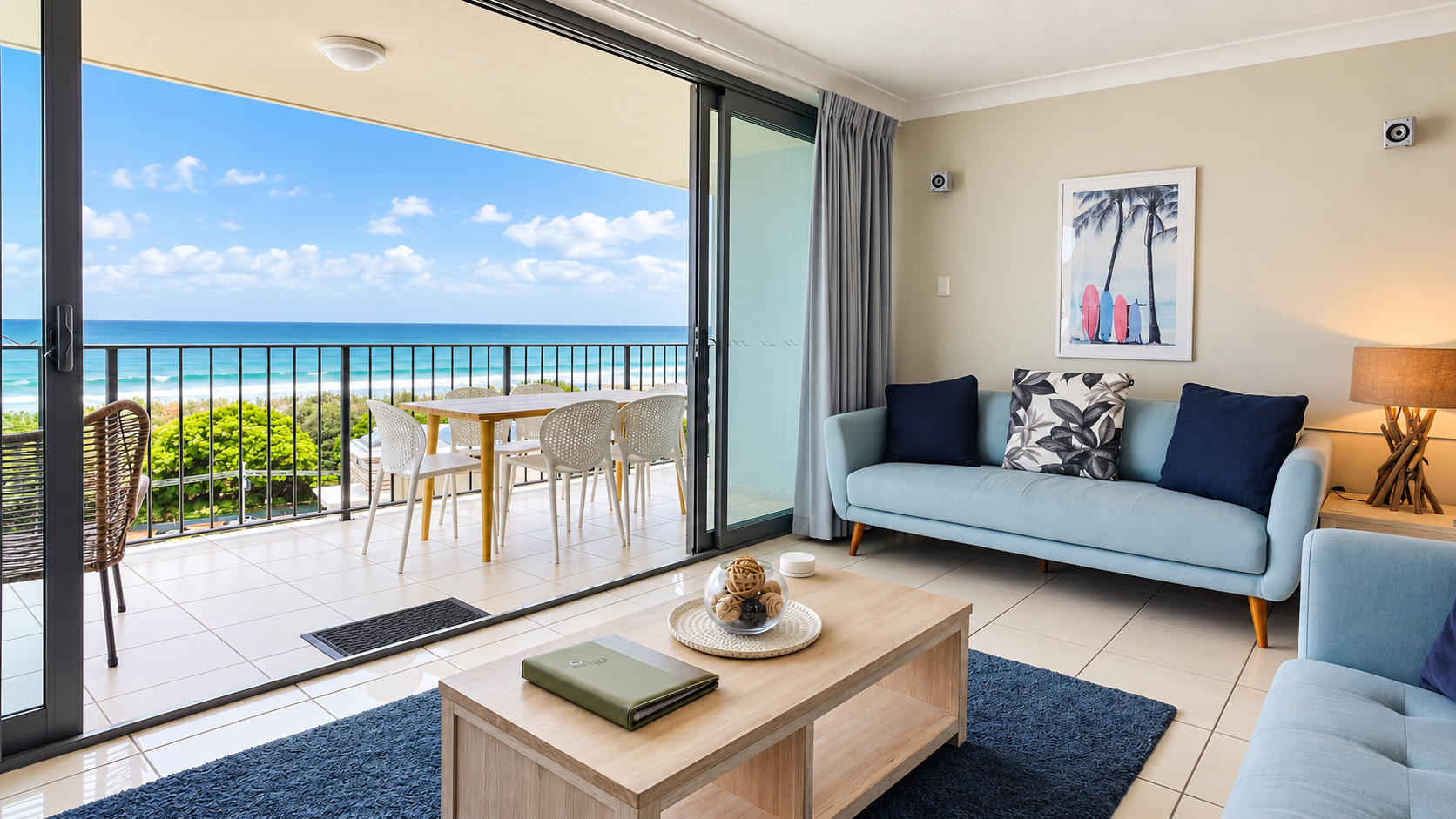 Modern living room with light blue sofas, dark blue and patterned cushions, wooden coffee table with decorative bowl, and sliding glass doors opening to a balcony with a dining table and chairs overlooking a beach and ocean under a partly cloudy blue sky.