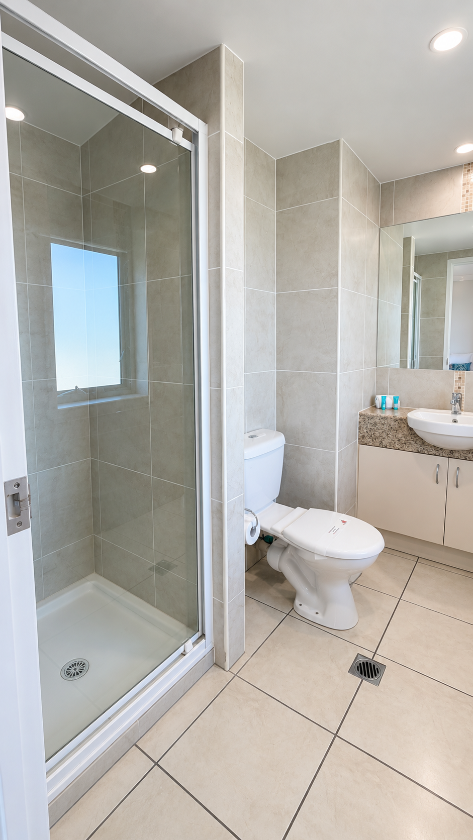 Bright modern bathroom with beige tiled walls and floor, featuring a glass shower enclosure with a window, a white toilet with a closed lid, and a vanity with granite countertop, a round white sink, a mirror, and toiletries.