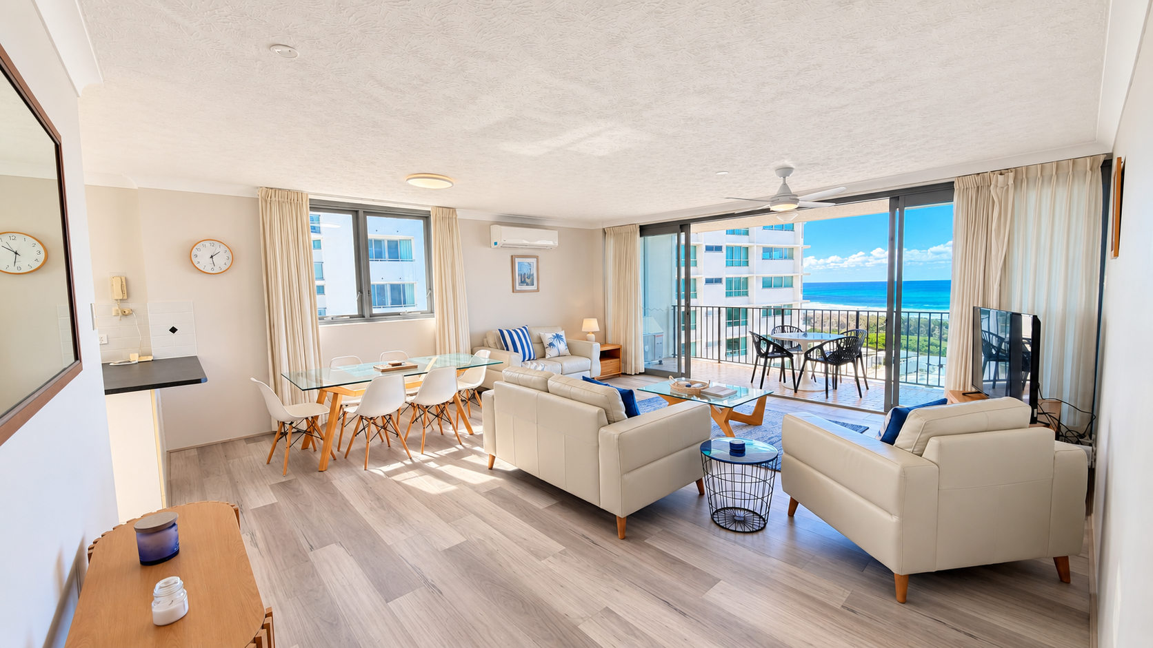 Bright, modern living room with light wood flooring, cream leather sofas with blue cushions, glass coffee and dining tables with white chairs, wall clock, and sliding glass doors opening to a balcony with ocean views and outdoor seating.