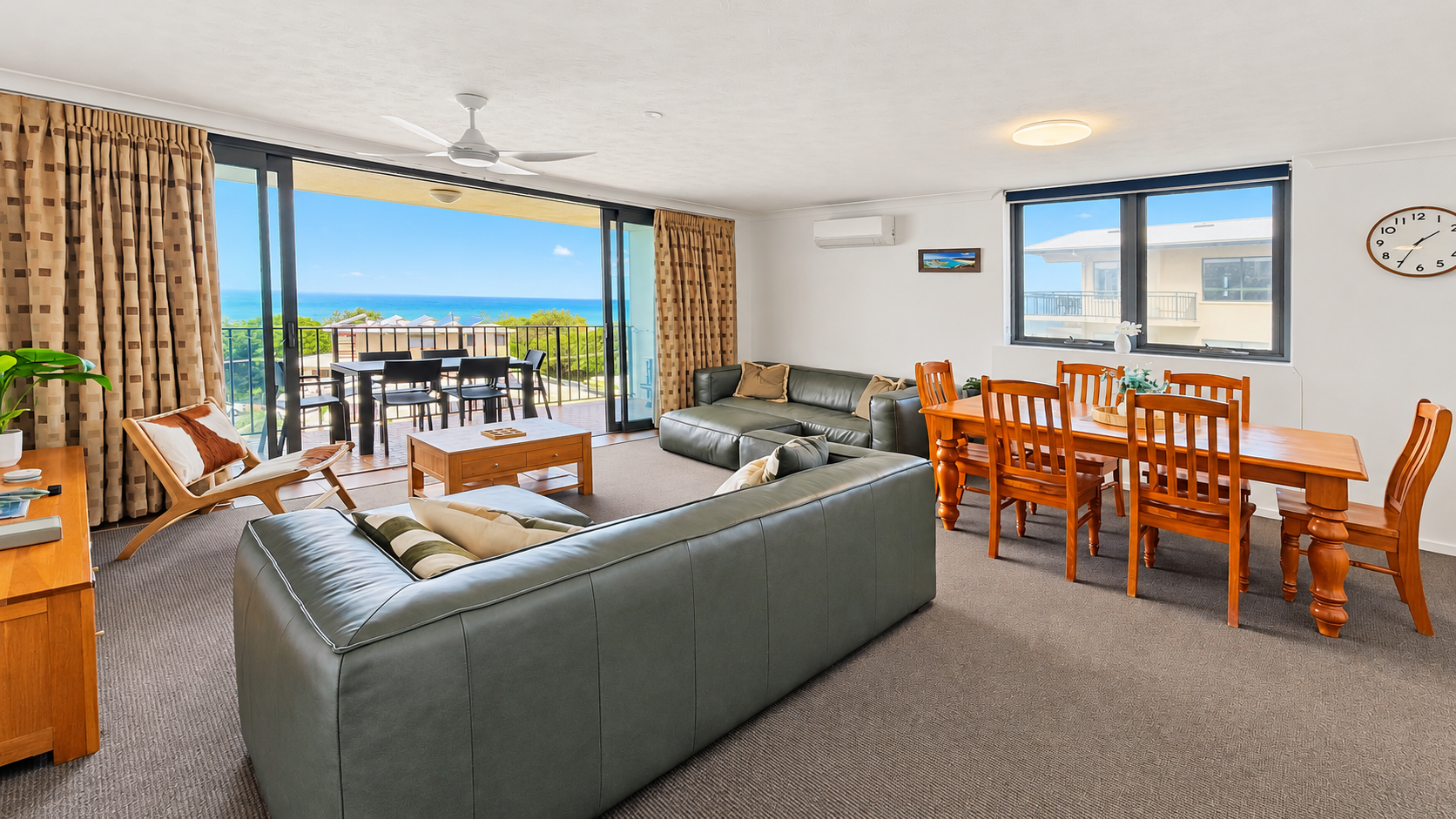 Bright living and dining area with two green leather sofas, wooden chair with patterned cushions, wooden coffee table, and a dining table with six chairs near a window; large sliding glass doors open to a balcony with outdoor table and chairs, overlooking the ocean and blue sky.