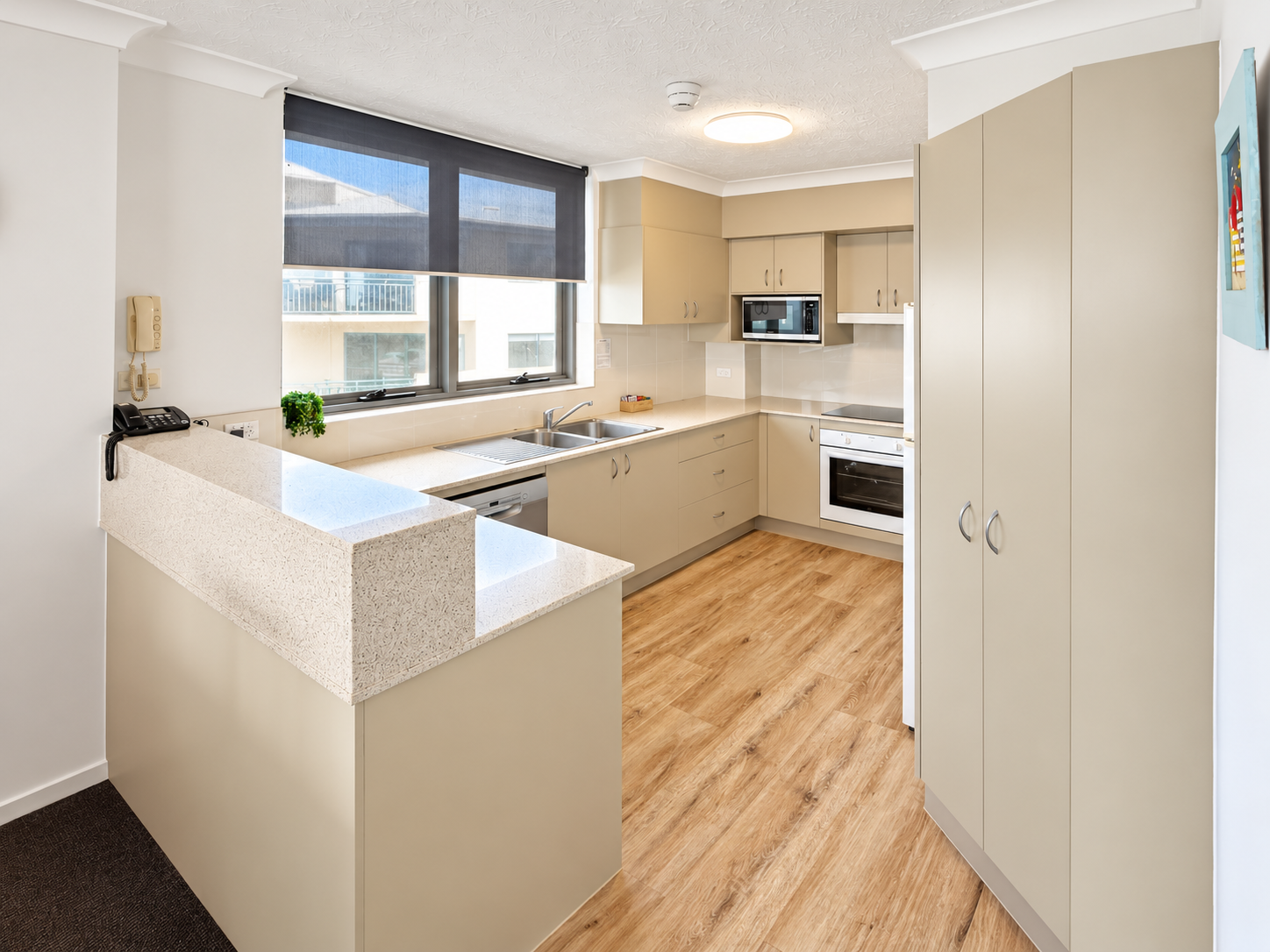 Modern kitchen with beige cabinets, light wood flooring, stone countertops, stainless steel sink, built-in oven, microwave, dishwasher, large window with partially lowered black roller blind, and a small green plant on the windowsill.