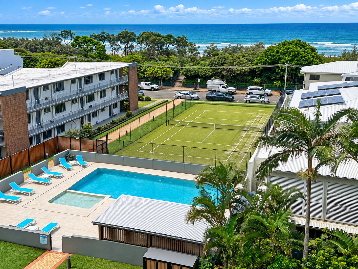 Aerial view of a residential area featuring a swimming pool with blue lounge chairs arranged around it, a small adjacent hot tub, a fenced tennis court with artificial grass, and nearby buildings with solar panels on the roof, set against a backdrop of trees and a blue ocean under a partly cloudy sky.