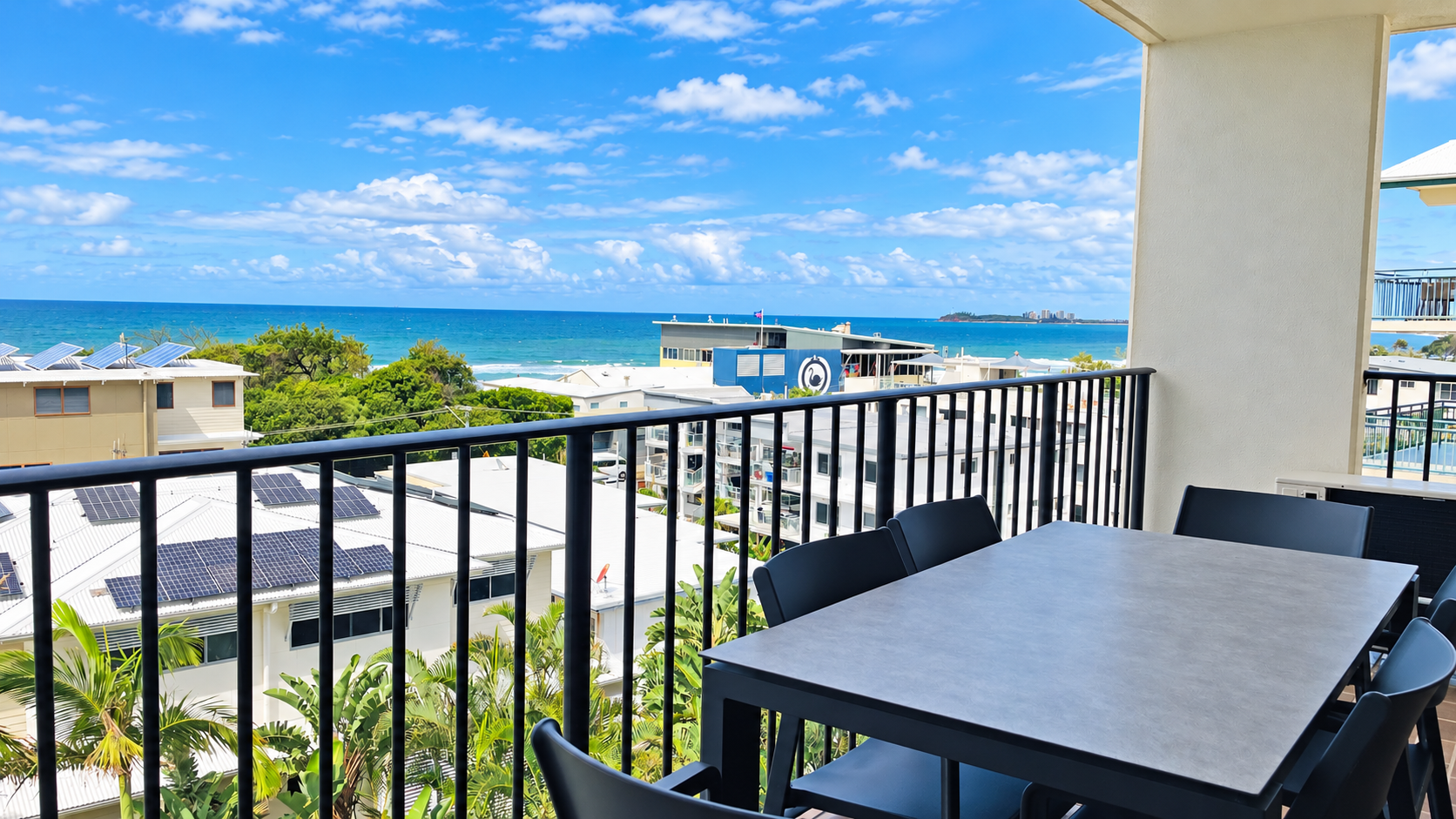 Balcony with black metal railing and a gray table surrounded by black chairs overlooking white buildings with solar panels, green trees, and a blue ocean under a partly cloudy sky.
