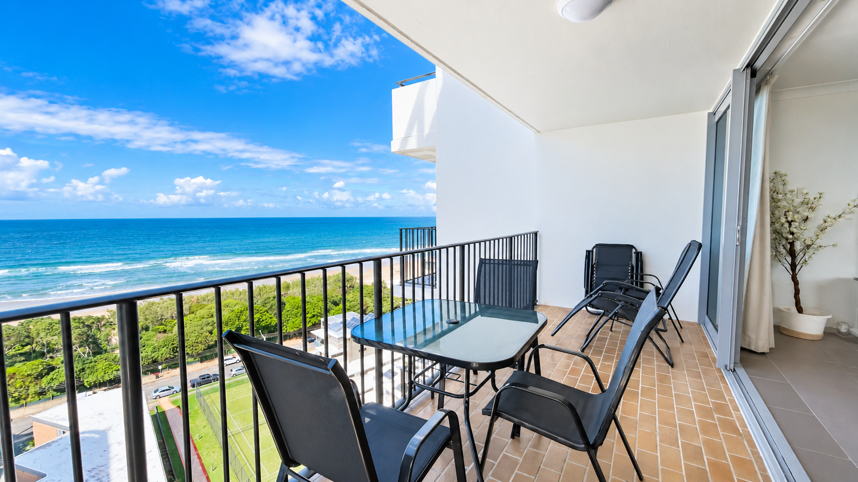Balcony with four black mesh chairs and a glass-top table overlooking a beach and ocean under a partly cloudy blue sky, with a green area and road below, and sliding glass doors leading inside.