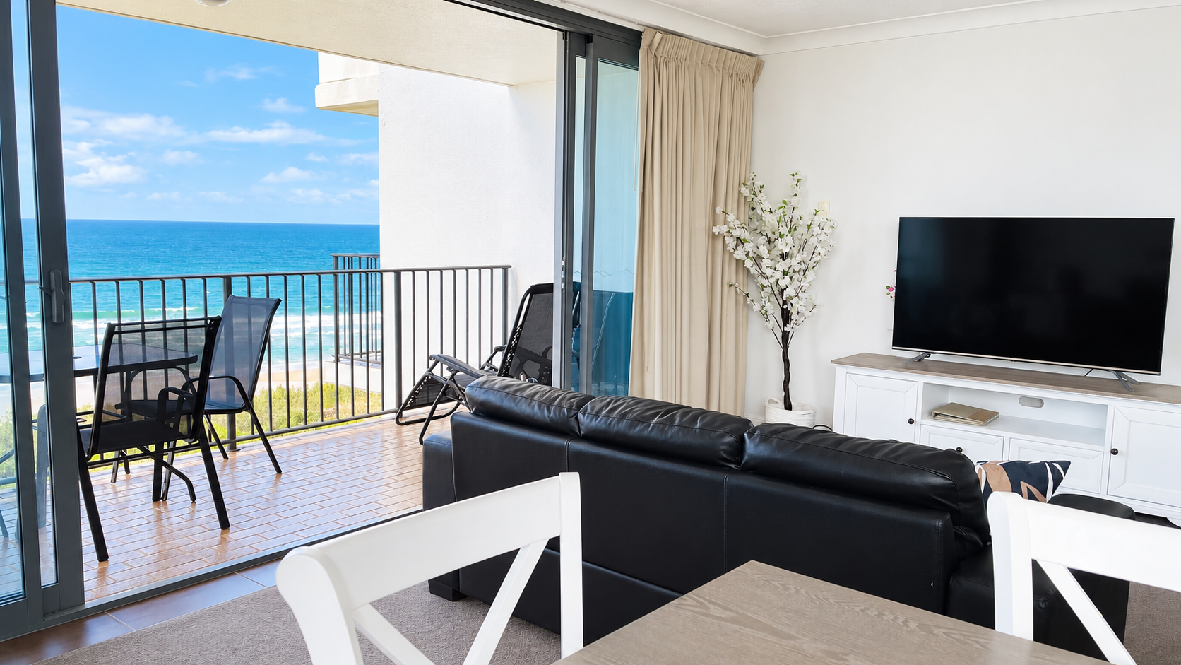 Living room with black leather couch facing a flat-screen TV on a white media console, beige curtains, artificial white flowers in a pot, and sliding glass doors opening to a balcony with black outdoor chairs and table overlooking the ocean.