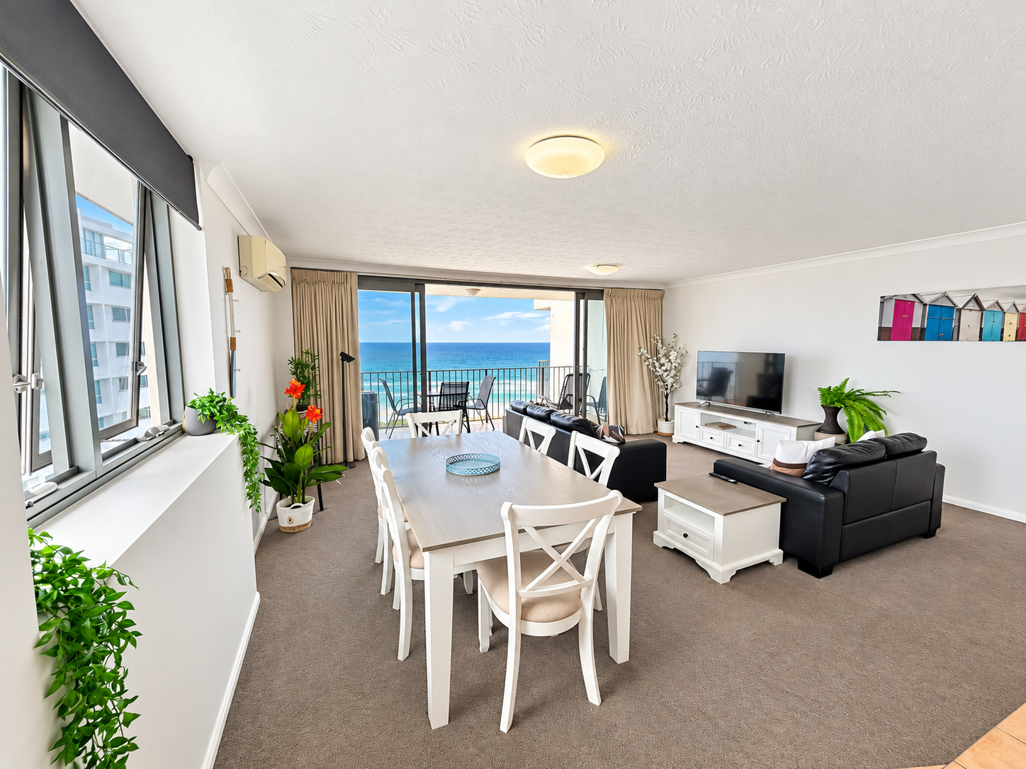 Bright lounge with beige carpet featuring a white dining table with six chairs, two black leather sofas facing a TV on a white stand, potted plants, and large sliding glass doors opening to a balcony with ocean view.