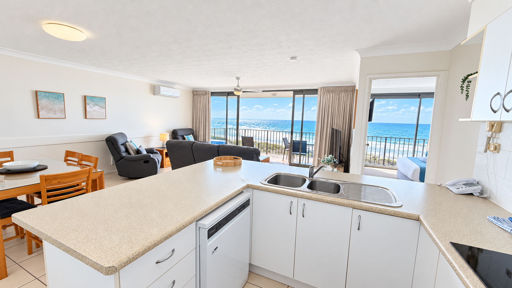 Bright open-plan kitchen and living area with beige countertops, white cabinets, a double sink, and wooden dining table with chairs. The living room has dark upholstered armchairs and a sofa facing a large flat-screen TV. Floor-to-ceiling sliding glass doors open to a balcony with outdoor seating and view of the ocean and blue sky. A bedroom with a bed is visible through a doorway to the right, also overlooking the ocean.