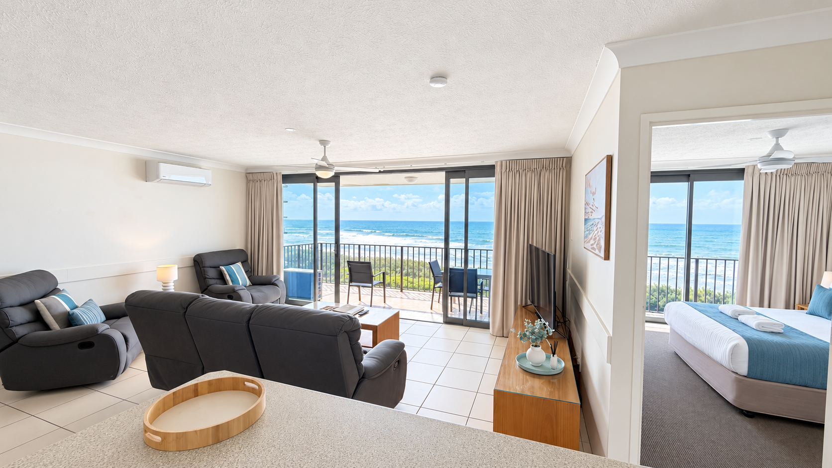 Bright living room with dark gray recliner sofas facing a wooden TV stand and large sliding glass doors opening to a balcony overlooking the ocean, with two chairs on the balcony and a bedroom visible to the right with a bed dressed in white and blue linens.