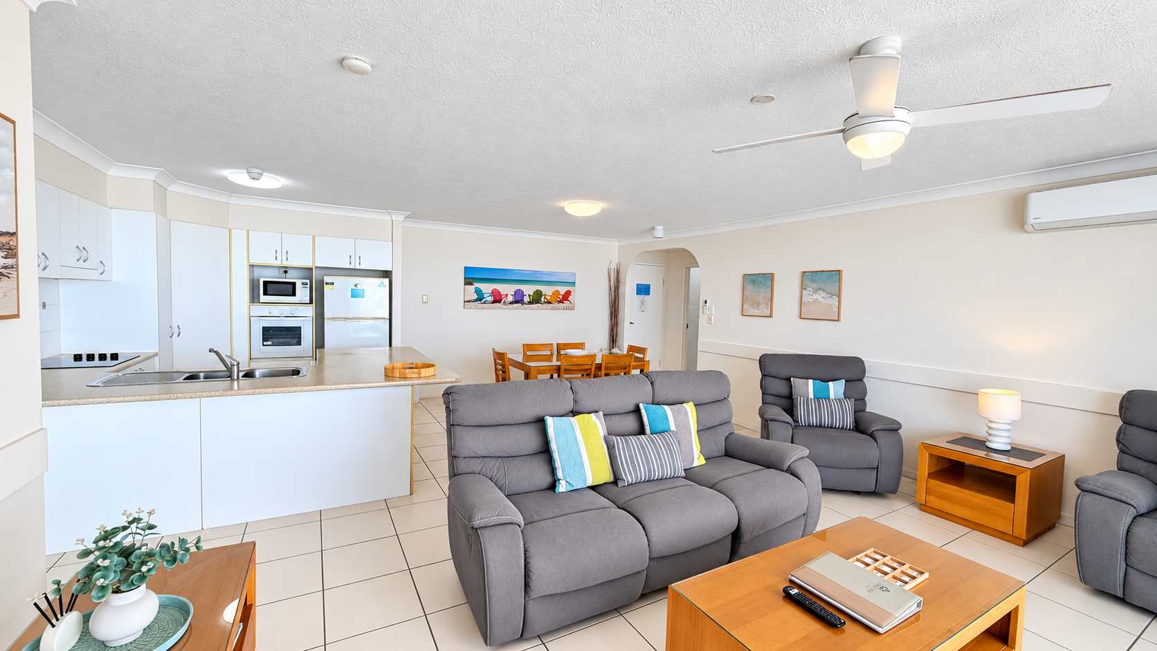 Spacious modern lounge with grey upholstered sofas and chairs with colorful striped cushions, wooden coffee and side tables, white tiled floor, open kitchen with white cabinets and appliances, and wooden dining table with six chairs against a wall with beach-themed artwork, ceiling fan, and air conditioner.