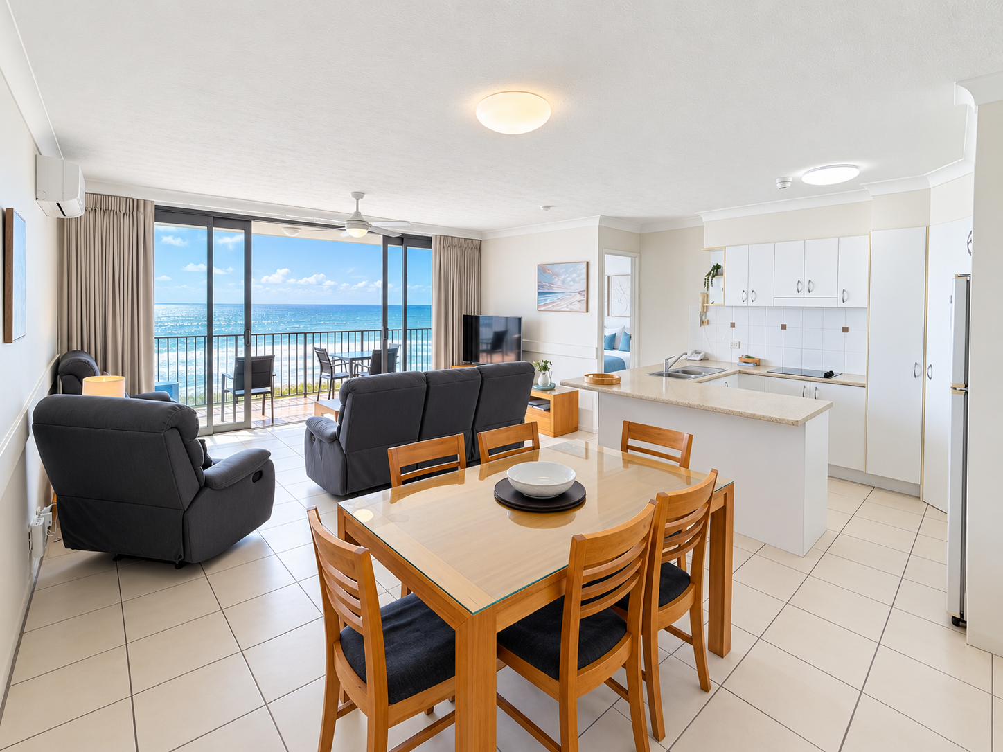 Bright open-plan living and kitchen area with a wooden dining table and six chairs, dark upholstered sofas, and a sliding glass door leading to a balcony with ocean views under a blue sky with clouds.