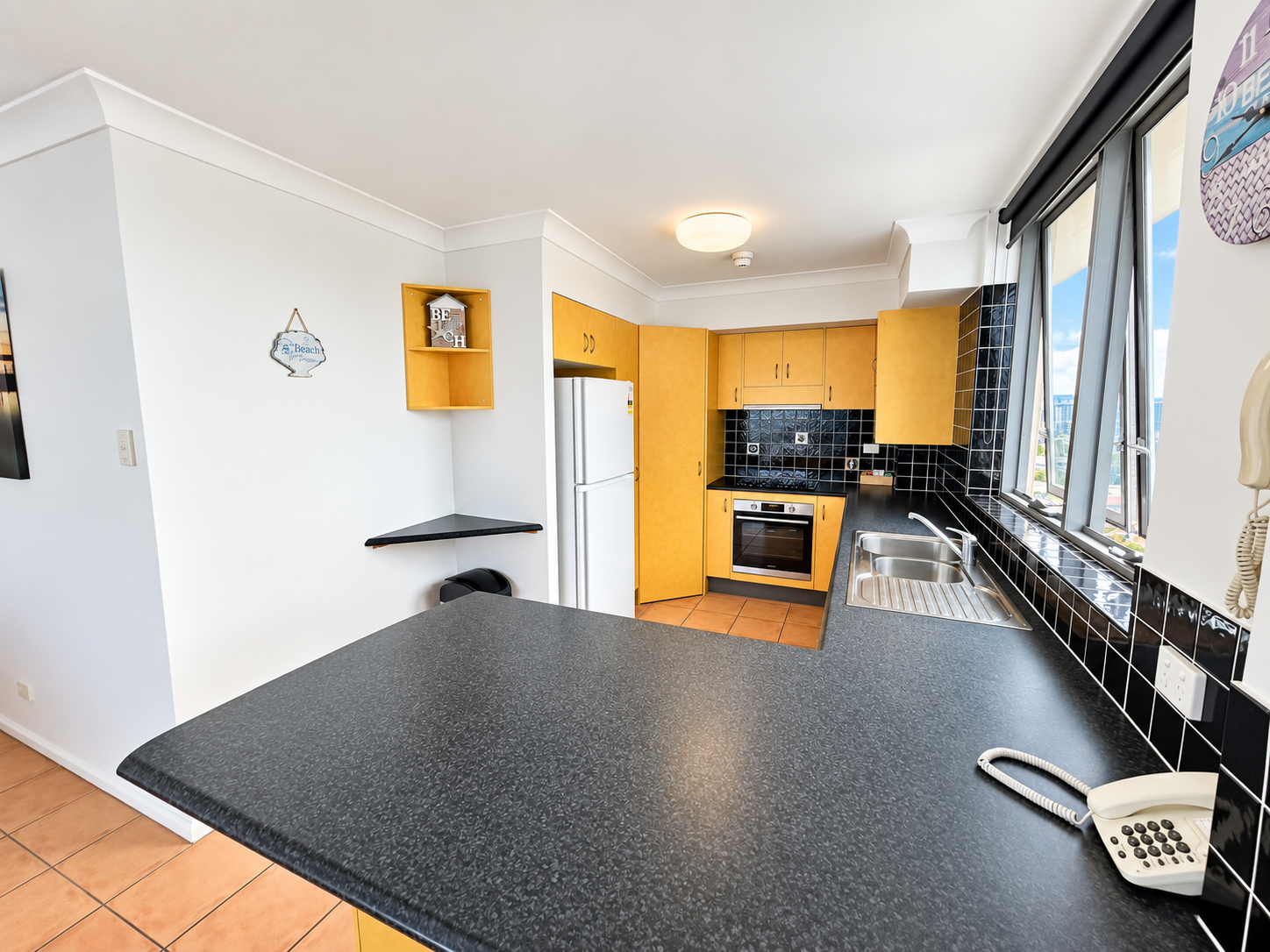 Modern kitchen with black countertops and black tile backsplash, light wood cabinets, a stainless steel oven, a double sink beneath large windows, and a white refrigerator in a corner.