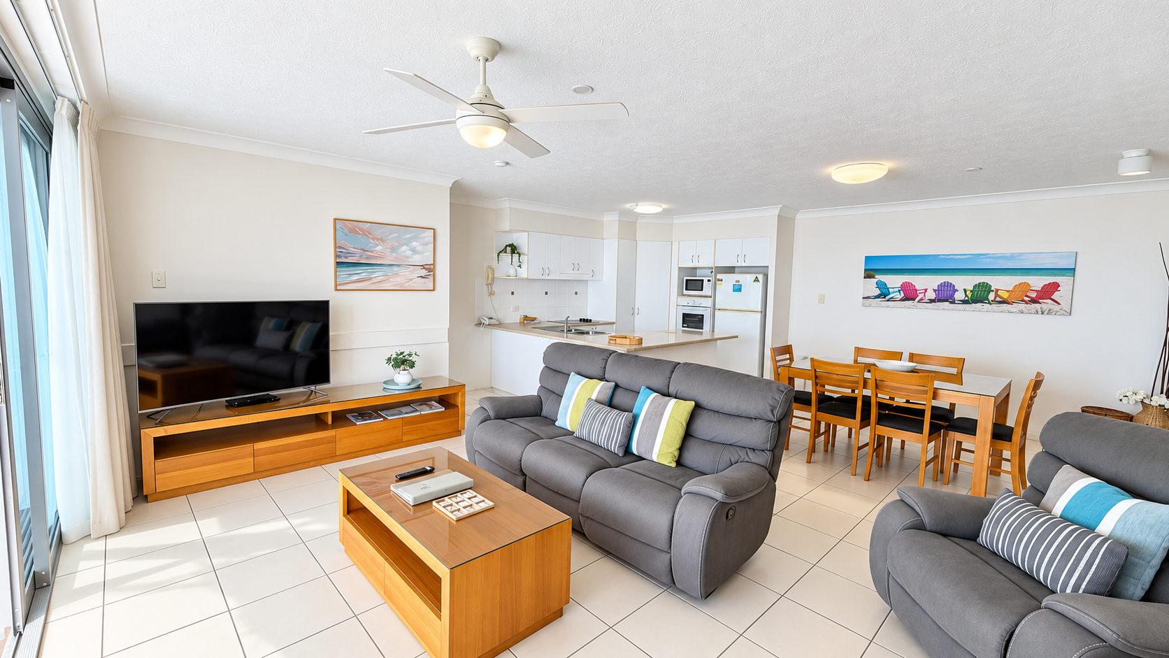 Bright open-plan living space with gray cushioned sofas, wooden coffee and TV tables, a flat-screen TV, dining table with six chairs, and a white kitchen with appliances, two beach-themed paintings on walls, and large windows with sheer curtains letting in sunlight.