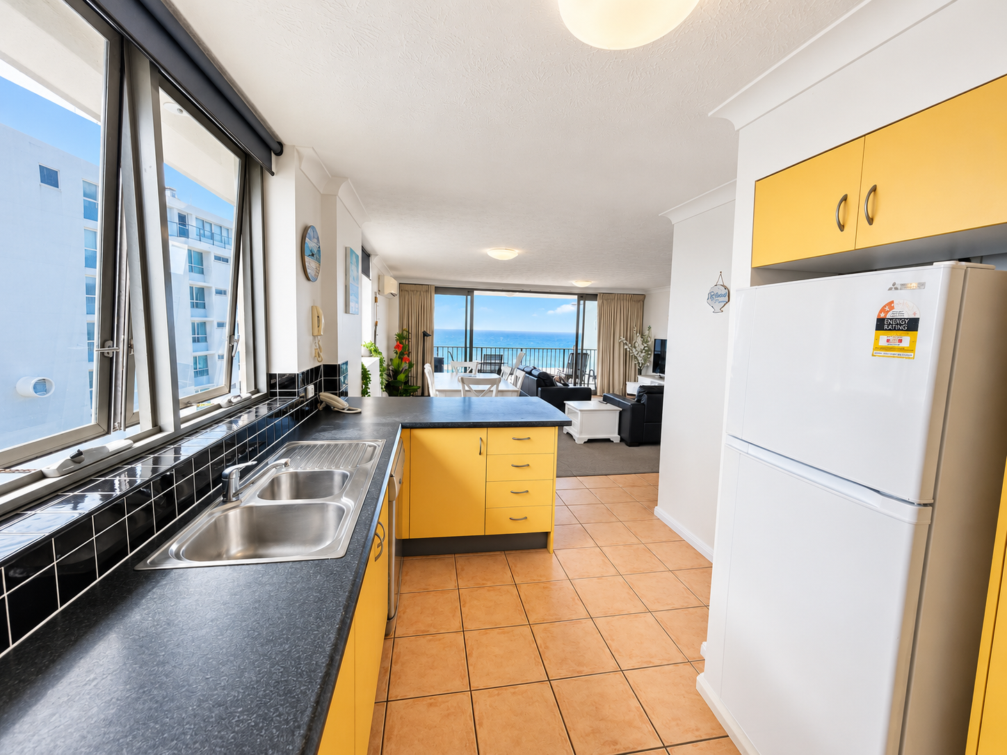 Modern kitchen with yellow cabinets, black countertop, double stainless steel sink, and a white refrigerator, opening into a living area with large windows showing an ocean view.