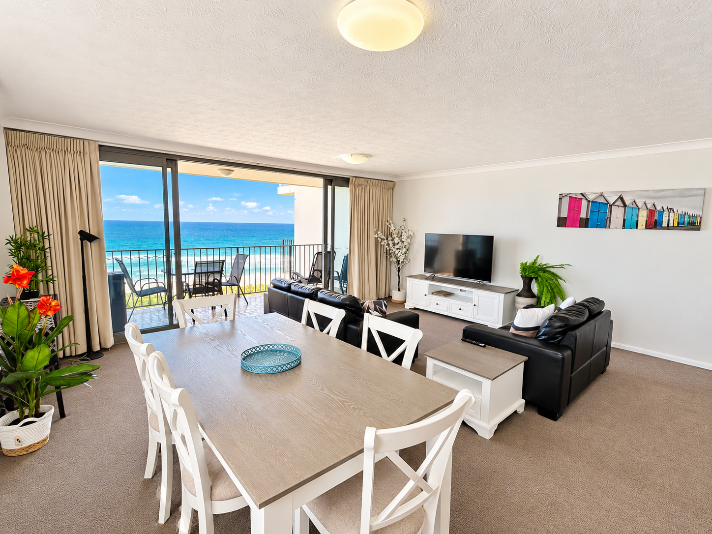 Bright dining and living area with a light wood table surrounded by white chairs, black leather sofas, a flat-screen TV on a white cabinet, plants, and sliding glass doors opening to a balcony with a table, chairs, and a view of the blue ocean and sky.