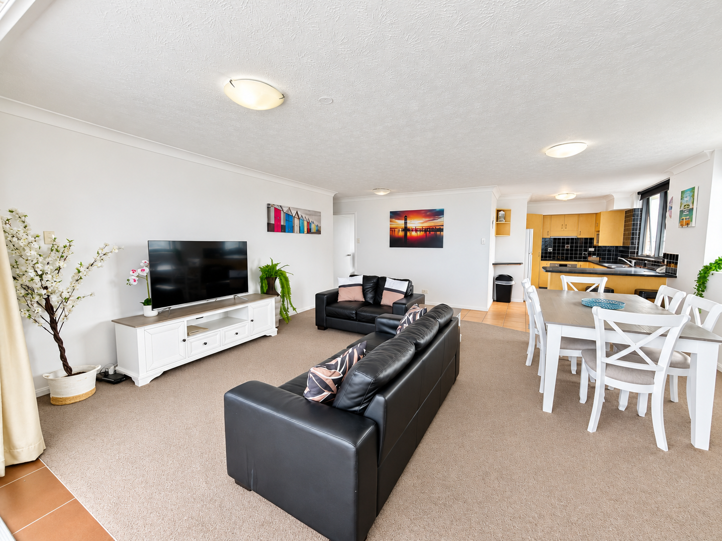 Modern open-plan living room with two black leather sofas facing a large flat-screen TV on a white TV stand, a white dining table with six chairs, and a kitchen with wooden cabinets and black tiled backsplash in the background.