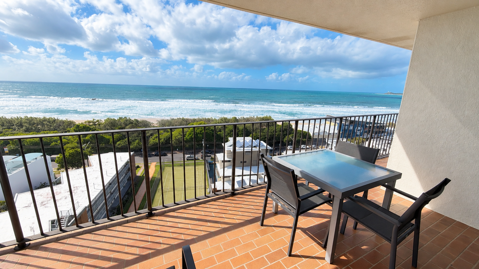 Balcony with a glass-top table and three black chairs overlooking a green tennis court, buildings, a tree line, sandy beach, and ocean under a partly cloudy sky.